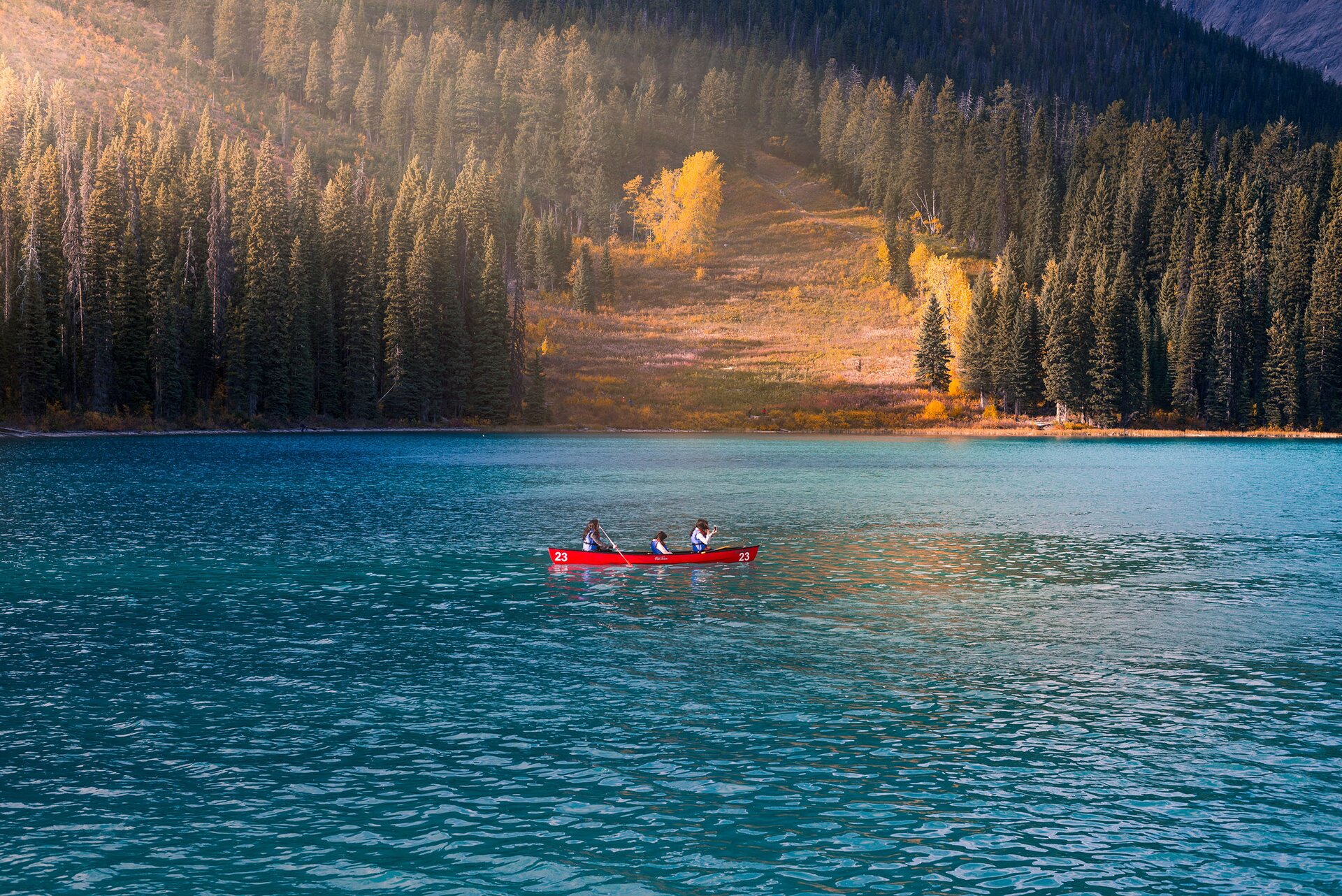 Zdjęcie przedstawia zbiornik wodny, na środku którego widoczny jest czerwony kajak. W kajaku widoczne są trzy osoby ubrane w niebieskie kapoki. Osoba siedząca na rufie trzyma wiosło zanurzone w wodzie. W głębi widoczne są schodzące do wody stoki porośnięte iglastymi drzewami i kilkoma liściastymi drzewami z żółtymi liśćmi.