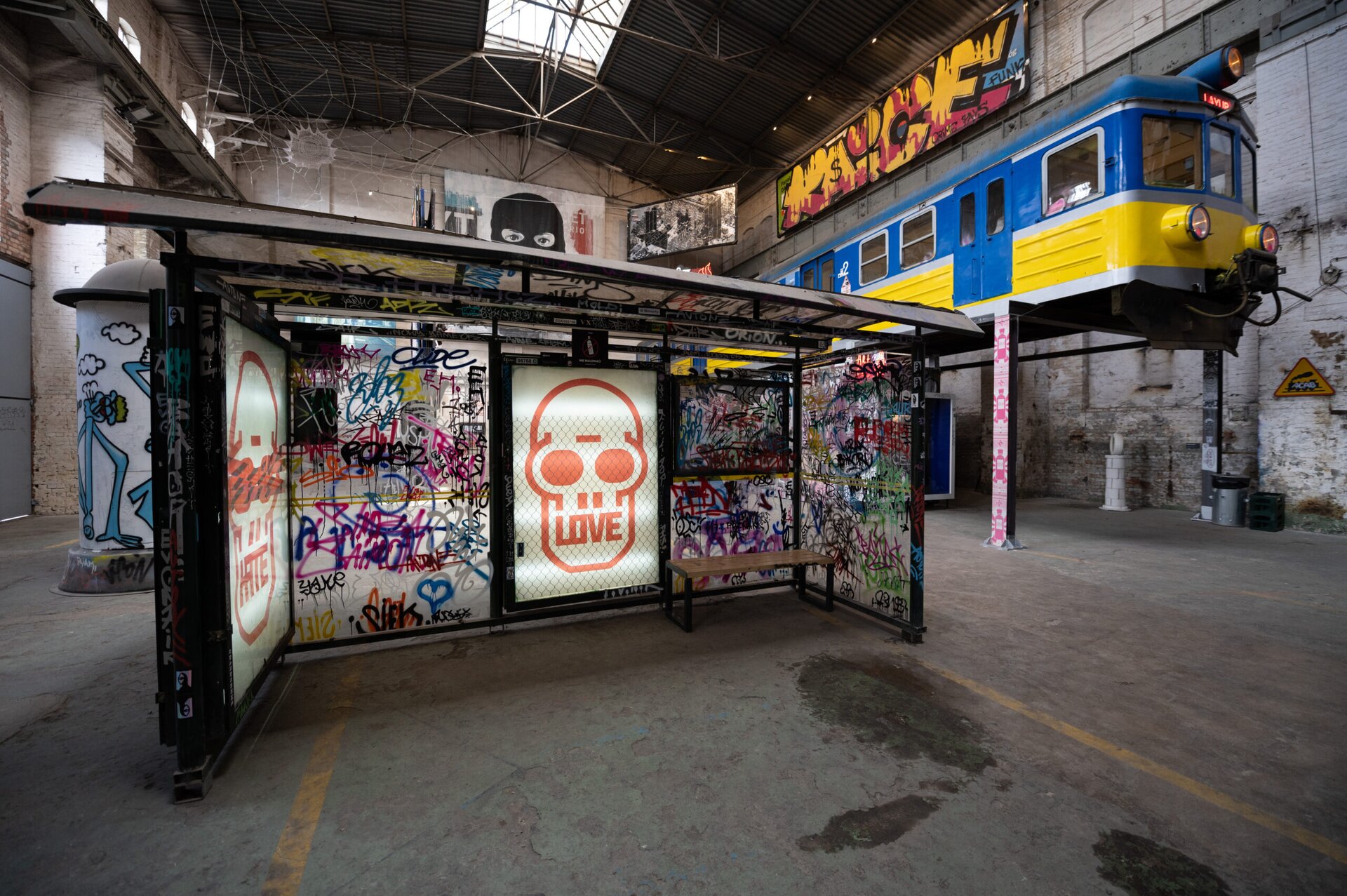 Na zdjęciu znajduje się wysokie pomieszczenie o jasnych, surowych ścianach i podłodze. Na pierwszym planie jest przystanek autobusowy, na którym jest wiele kolorowych graffiti [czytaj: grafiti]. Za nim, po lewej stronie, jest biało‑szary słup ogłoszeniowy. Na słupie są kolorowe malunki. Na drugim planie, po prawej stronie, na podwyższeniu jest wagon pociągu. Jest on w kolorze niebiesko‑żółtym. W tle są duże, kolorowe graffiti [czytaj: grafiti].