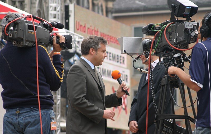 Zdjęcie przedstawia reportera telewizyjnego z mikrofonem, który stoi przed operatorami z kamerami. Przeprowadza wywiad z innym mężczyzną. Obydwaj mężczyźni są ubrani w garnitury.