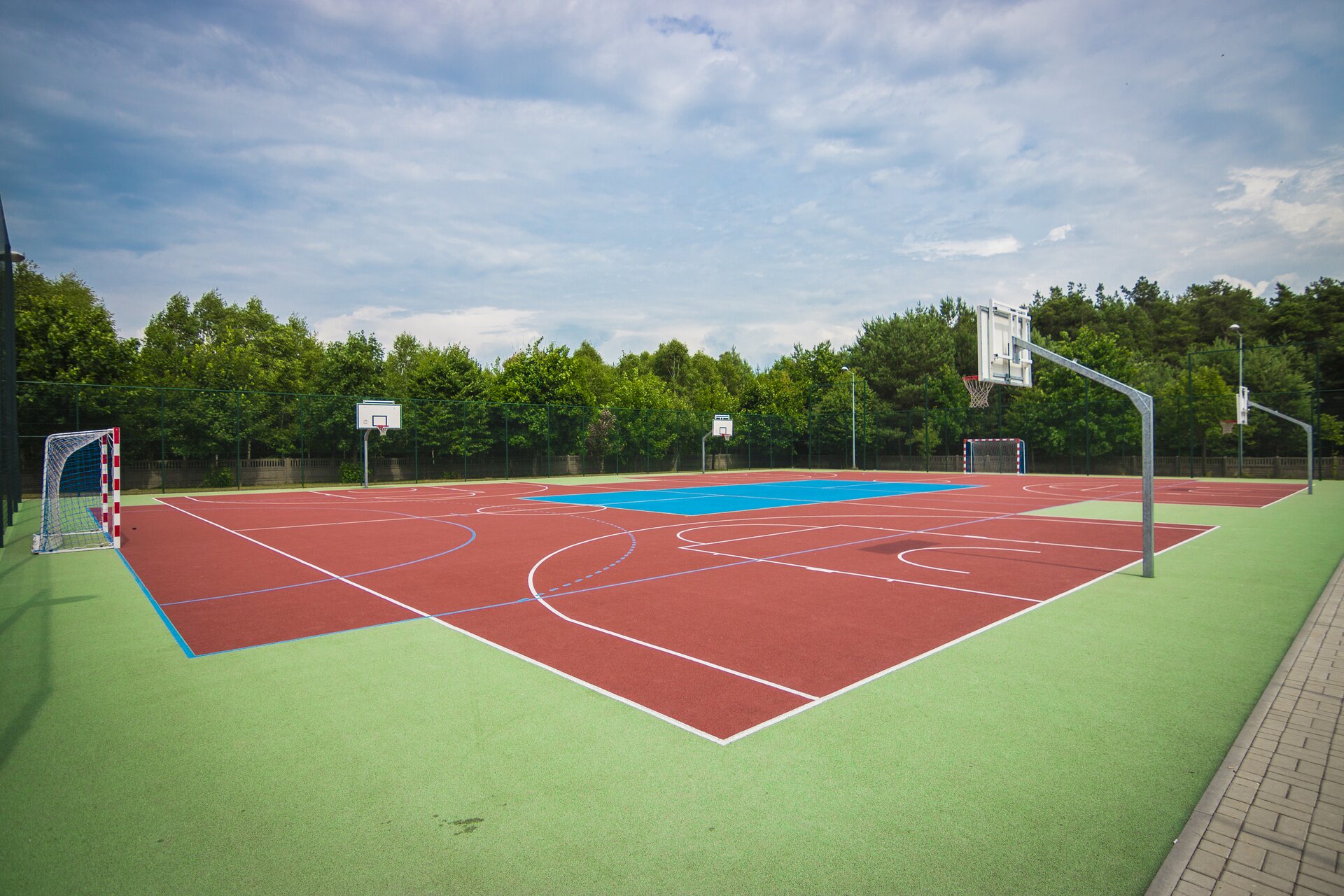 Zdjęcie przedstawiające boisko szkolne. Składa się z czterech koszów do koszykówki i dwóch bramek do piłki nożnej. Wokół rosną drzewa.