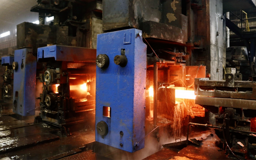 Obrazek jest fotografią, która przedstawia walcarki hutnicze na hali produkcyjnej. Są to maszyny umieszczone kolejno po sobie, składające się z dwóch stalowych walców, przez które przeprowadzone są rozgrzane do czerwoności elementy metalowe. Metalowe elementy przechodzą przez kolejne walcarki zmieniając swój kształt w zależności od wielkości walców oraz ich rozchylenia.