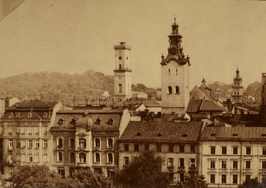 Fotografia przedstawia kilkukondygnacyjne kamienice. Górują nad nimi dwie wieże. W tle porośnięte drzewami pagórki.