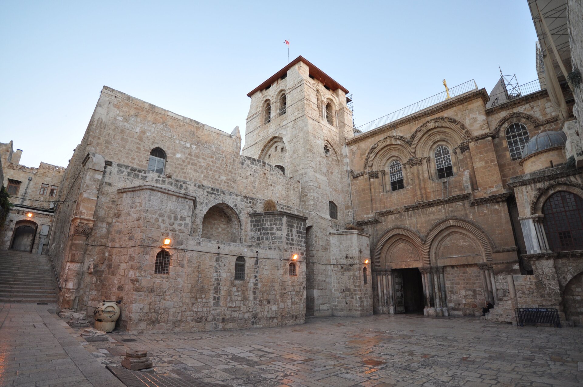 Ilustracja przedstawia zdjęcie współczesnej bazyliki Grobu Pańskiego w Jerozolimie. Na fotografii ukazany jest zabytkowy kościół z wieżą o podwójnych oknach na planie kwadratu. Po prawej stronie wieży, w dwukondygnacyjnej fasadzie znajdują się dwa portale zwieńczone kilkoma rzędami ostrych łuków podtrzymywanych trzema kolumnami po obu stronach drzwi. Wejście po prawej stronie jest zamurowane. Na drugiej kondygnacji znajdują się dwa ostrołukowe okna. Na środku wielostopniowej fasady, po lewej stronie wieży znajduje się ślepa ostrołukowa wnęka. Poniżej natomiast mieszczą się trzy małe okienka. Bazylika zbudowana jest z czerwonej cegły. W tle ukazane są schody prowadzące do wejścia do kamienicy Starego Miasta Jerozolimy.