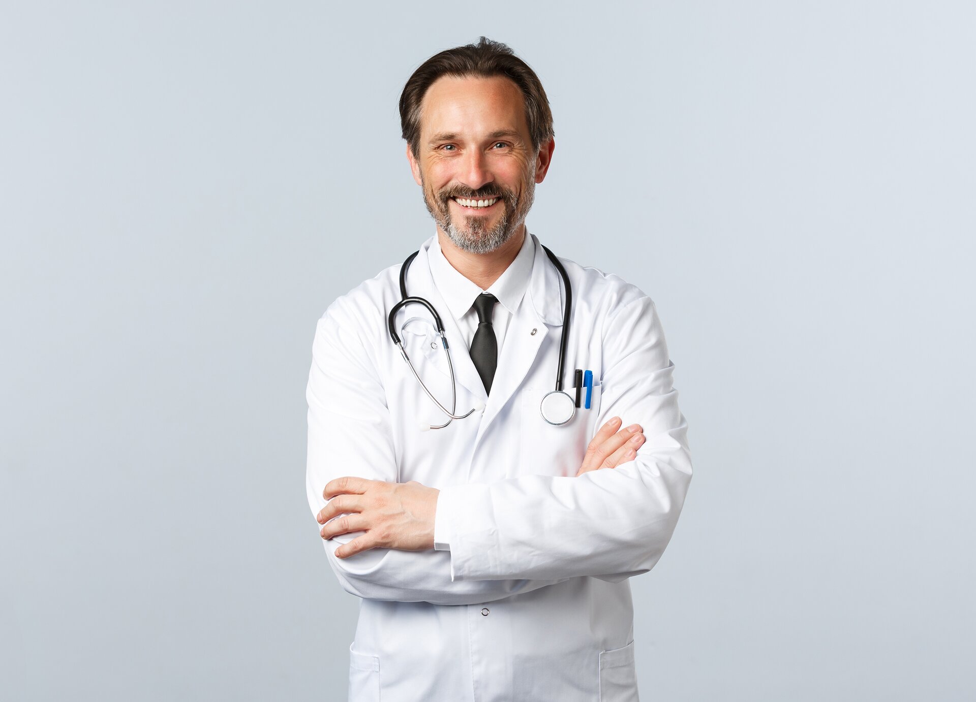 Zdjęcie przedstawia stojącego mężczyznę w średnim wieku. Mężczyzna jest ubrany w biały fartuch lekarski, ma na szyi przewieszony stetoskop, a w kieszonce fartucha, znajdującej się po lewej stronie na wysokości klatki piersiowej, ma włożone dwa długopisy. Mężczyzna uśmiecha się i ma ręce splecione na wysokości brzucha.