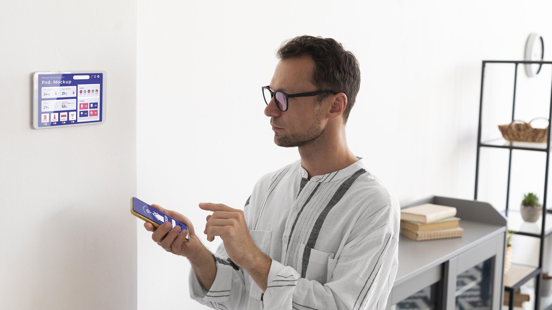 Zdjęcie przedstawia mężczyznę stojącego przy ścianie, na której znajduje się panel sterowania inteligentnym domem. Mężczyzna trzyma w dłoniach telefon i łączy się z panelem sterowania. 
