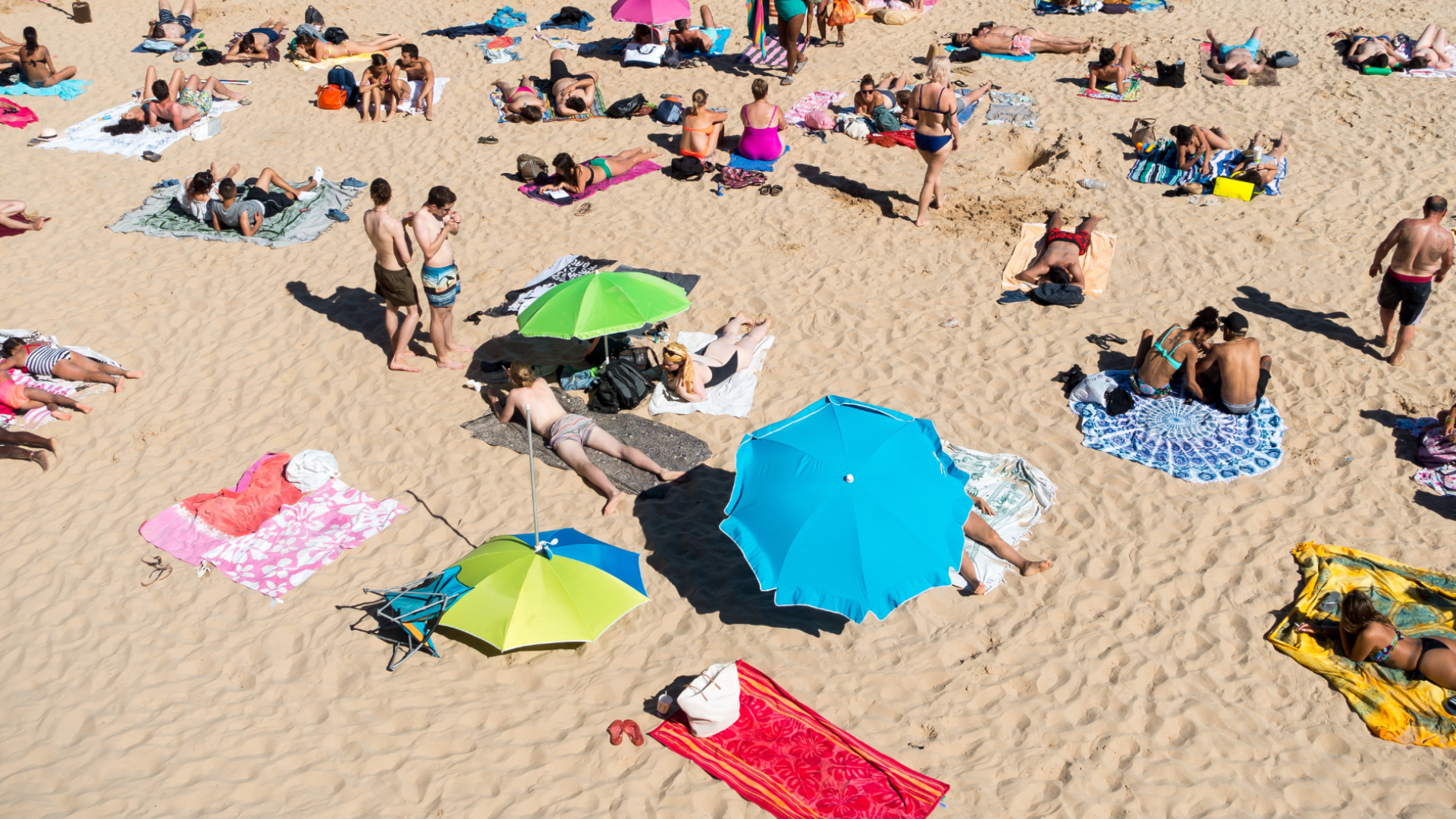 Zdjęcie zrobione z góry przedstawia ludzi na plaży. Na zdjęciu znajdują się ludzie, którzy leżą, albo siedzą na ręcznikach. Niektórzy chronią się przed słońcem pod dużymi parasolami.
