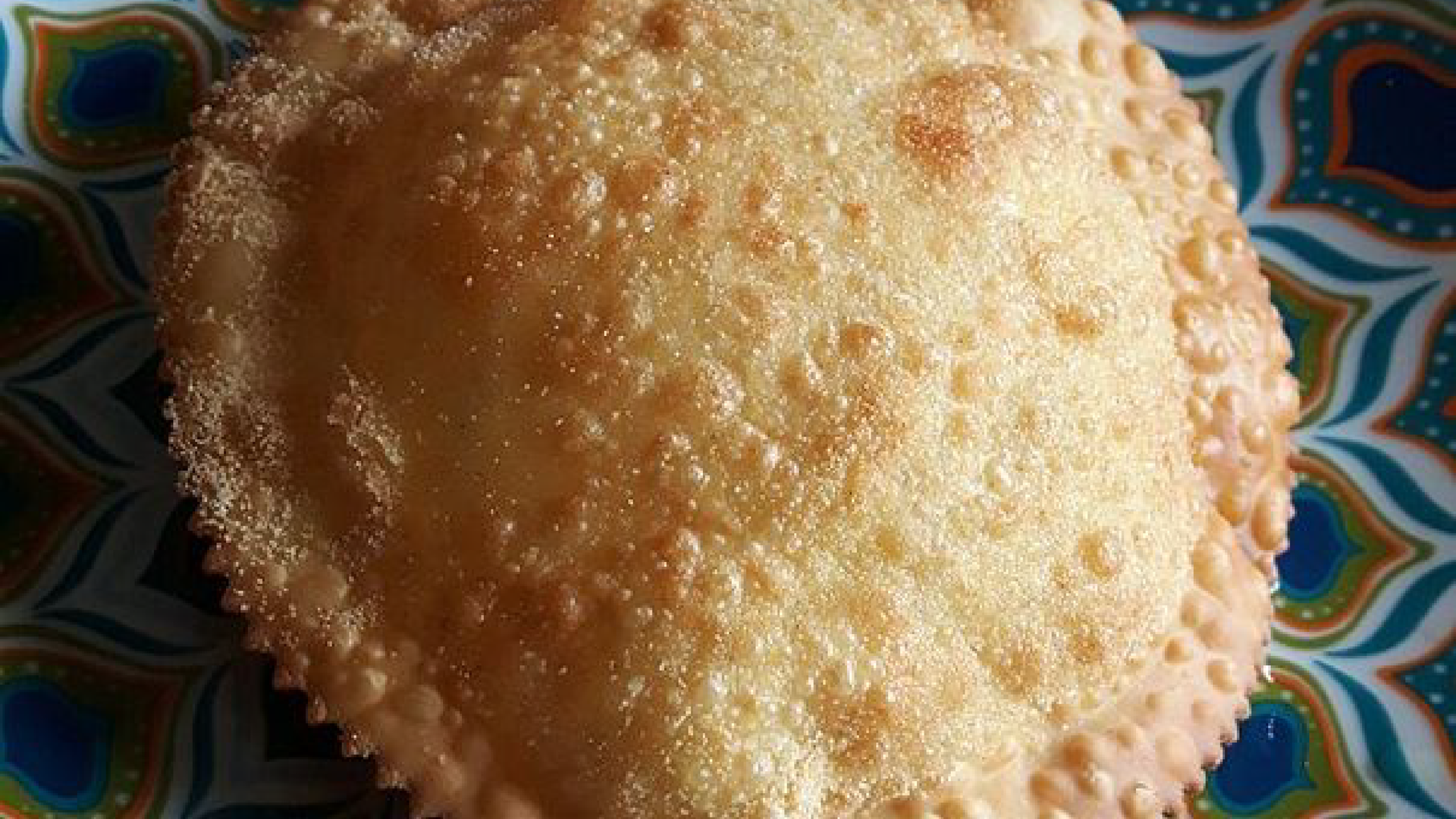 Zdjęcie przedstawia sardyński deser Seada, który jest smażony na głębokim oleju, co skutkuje jego szorstką powierzchnią pokrytą bąbelkami.