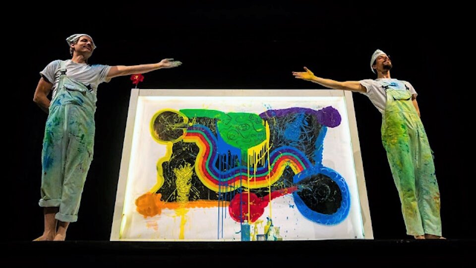 Zdjęcie przedstawia ciemną scenę. Pośrodku stoją dwaj mężczyźni, wskazując na siebie wyciągniętymi w bok rękami. Pomiędzy nimi znajduje się duży obraz sięgający im do ramion. Malarze w ubrudzonych farbą kombinezonach i śmiesznych czapeczkach z dumą prezentują swoje dzieło. Obraz to abstrakcja swobodnie malowana, z zaciekami spływających farb, przez środek płótna wije się wielobarwna rzeka. Zdjęcie zostało wykonane z dołu z poziomu teatralnej widowni.