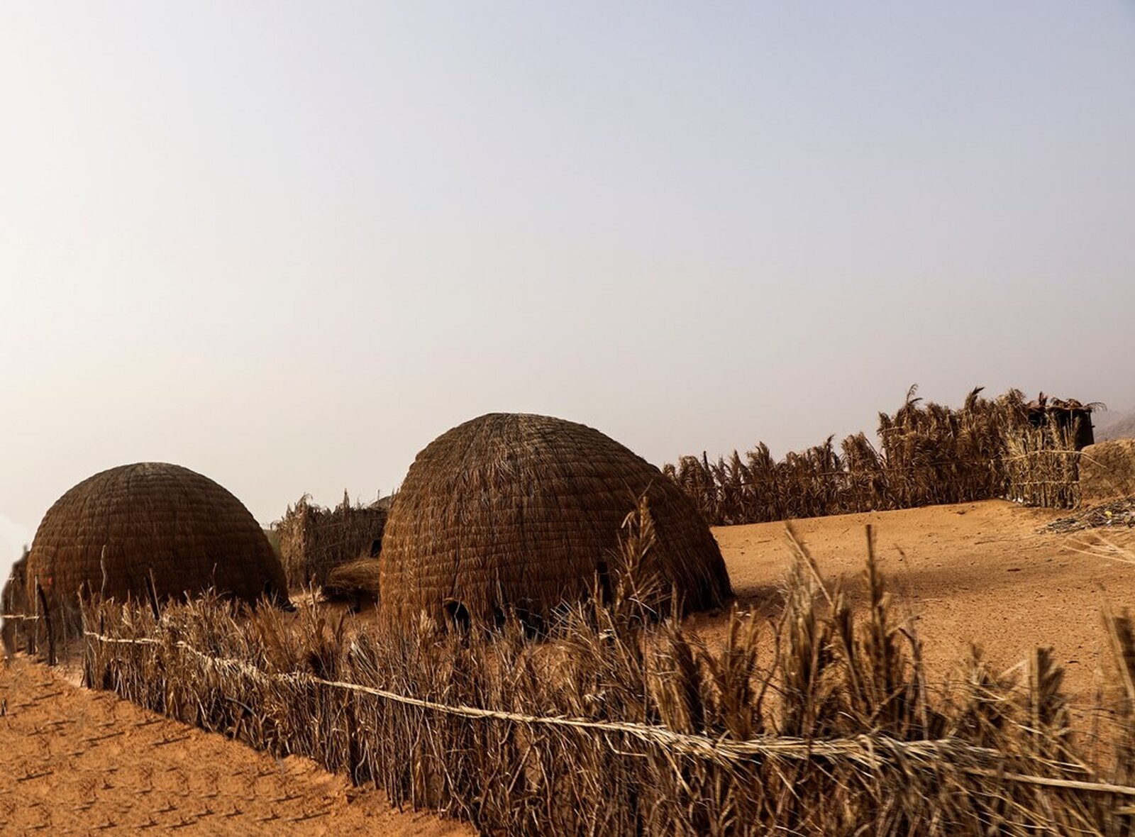 Zdjęcie przedstawia chaty tikkit otoczone zeribą. Domki przypominające kopulaste lepianki są otoczone niewielkim płotem zrobionym z gęsto plecionej trawy. Dookoła suchy, pomarańczowy piasek. 