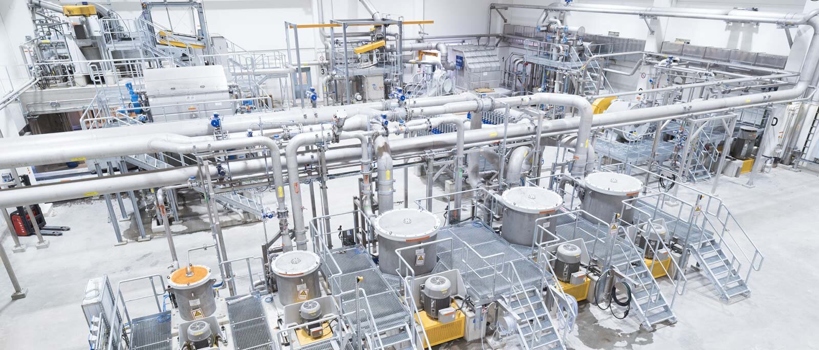 Zdjęcie ukazuje dział przygotowania mas włóknistych w zakładzie papierniczym. Na kilku podestach, do których prowadzą metalowe schodki, umieszczono różnej wielkości kadzie. Nad nimi znajduje się system rur. W tle widoczna jest pozostała część zakładu.