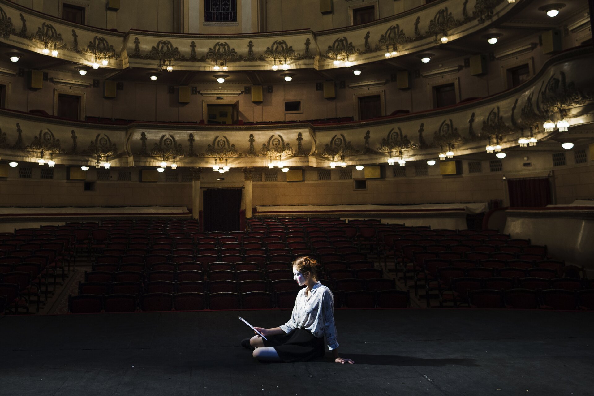 Zdjęcie przedstawia salę teatralną. Na scenie z przodu siedzi aktorka, trzymając kartki z tekstem w dłoniach. Przed sceną rozciągają się rzędy foteli. Wokół widowni widoczne są pięknie oświetlone balkony, które mają kształt półokręgów, jeden na dolnym poziomie i drugi na górnym.