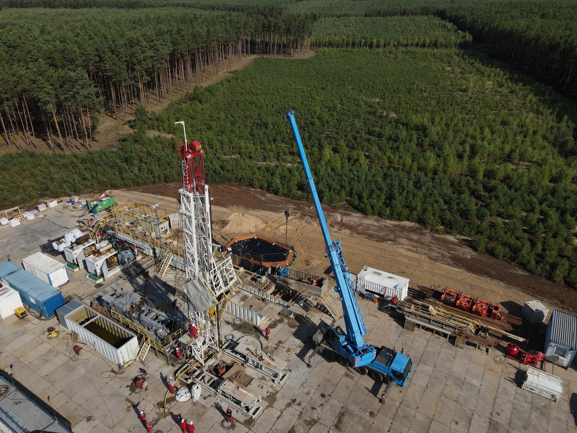 Na zdjęciu widać maszt wiertniczy znajdujący się na placu wiertniczym widoczny z góry.Plac wiertniczy widoczny jest z lotu ptaka. Ma kształt prostokąta. Podstawa placu jest wyłożona betonowymi płytami. W centralnym punkcie placu znajduje się maszt wiertniczy. Maszt jest w kształcie ostrosłupa ściętego. Maszt znajduje się pomiędzy białymi kontenerami. Po prawej stronie masztu widoczny jest niebieski dźwig. Przed masztem stoją ludzie ubrani w czerwone stroje ochronne i białe kaski. Podest masztu jest w czasie budowy.W tle zdjęcia widać tereny leśne.