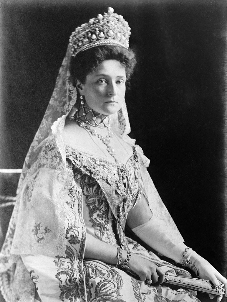 Fotografia przedstawia kobietę ubraną w eleganckie szaty, na szyi wisi ozdabiany naszyjnik, na głowie ma koronę wysadzaną drogimi kamieniami. Kobieta siedzi, ręce ma złożone na udach, w jednej z nich trzyma wachlarz.