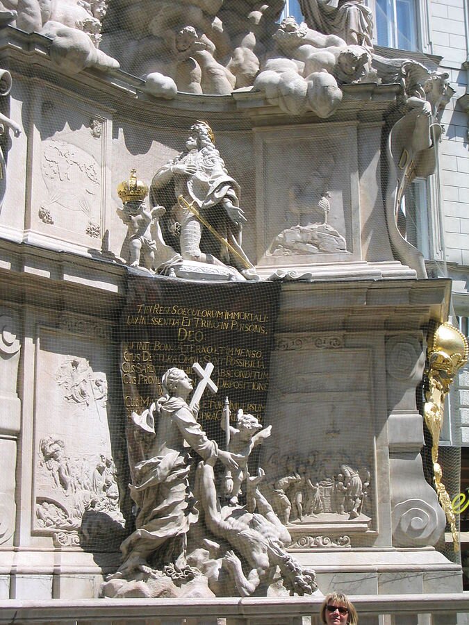 Zdjęcie przedstawia fragment kolumny morowej. Widnieją na nim klęczący i modlący się cesarz Leopold I, a poniżej postać kobiety symbolizującej wiarę, która u stóp ma umierającą postać zarazy, dobijaną przez małego cherubina.