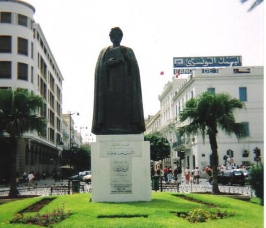 Na zdjęciu znajduje się pomnik Ibn Chalduna.  Stoi on na podeście na którym widnieją arabskie napisy. Położony jest w małym parku w centrum miasta przy ruchliwej ulicy. W tle znajdują się budynki.