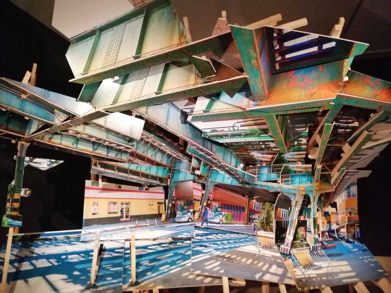 Zdjęcie przedstawia instalację wielkich rozmiarów, zbudowaną ze stalowych części pociągów, torów i kolejowych konstrukcji. Światło przechodzi przez ażurowe elementy, tworząc na podłodze wrażenie świetlnych torów. Pod zawieszoną instalacją znajdują się ogromne zdjęcia ułożone po bokach. Przedstawiają fragmenty dworców kolejowych. 