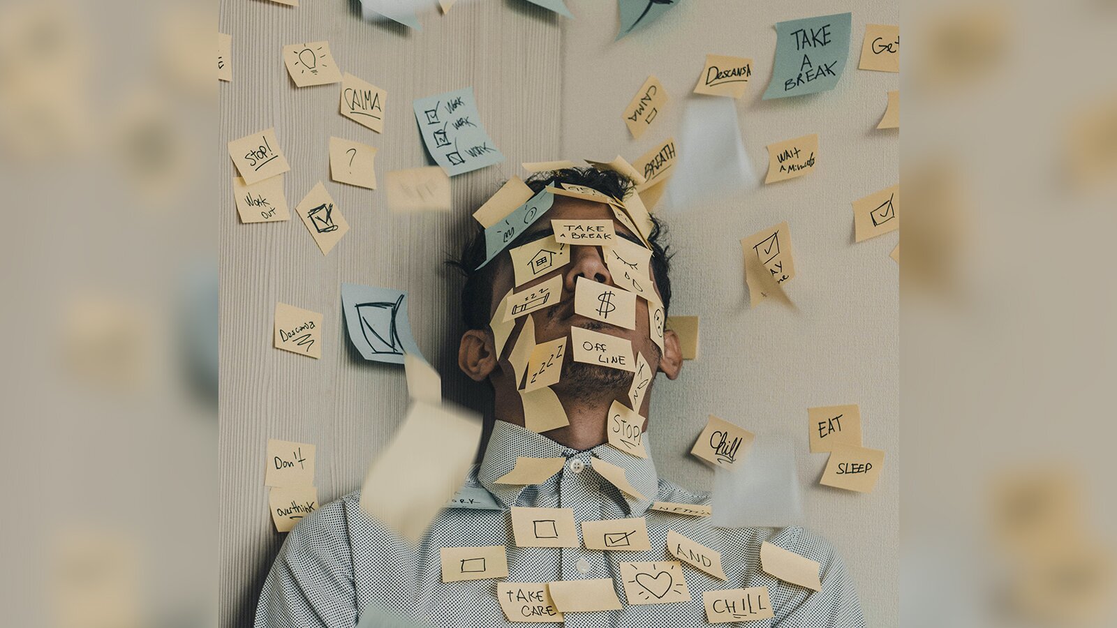 Zdjęcie przedstawia mężczyznę, w ujęciu od klatki piersiowej w górę, stojącego w rogu pomieszczenia. Ściany oraz mężczyzna są obklejeni karteczkami samoprzylepnymi w kolorze niebieskim i żółtym. Na karteczkach widoczne są różne symbole i słowa. Widoczne są między innymi: serca, puste kwadraty, żarówka, dom, symbol dolara amerykańskiego, kwadraty ze znakiem zatwierdzającym, tak zwanym ptaszkiem. Słowa zapisane na karteczkach to:: Stop; Work out; Don't overthink; Take care; And; Chill; Off line; Eat; Sleep; Breath; Take a break.