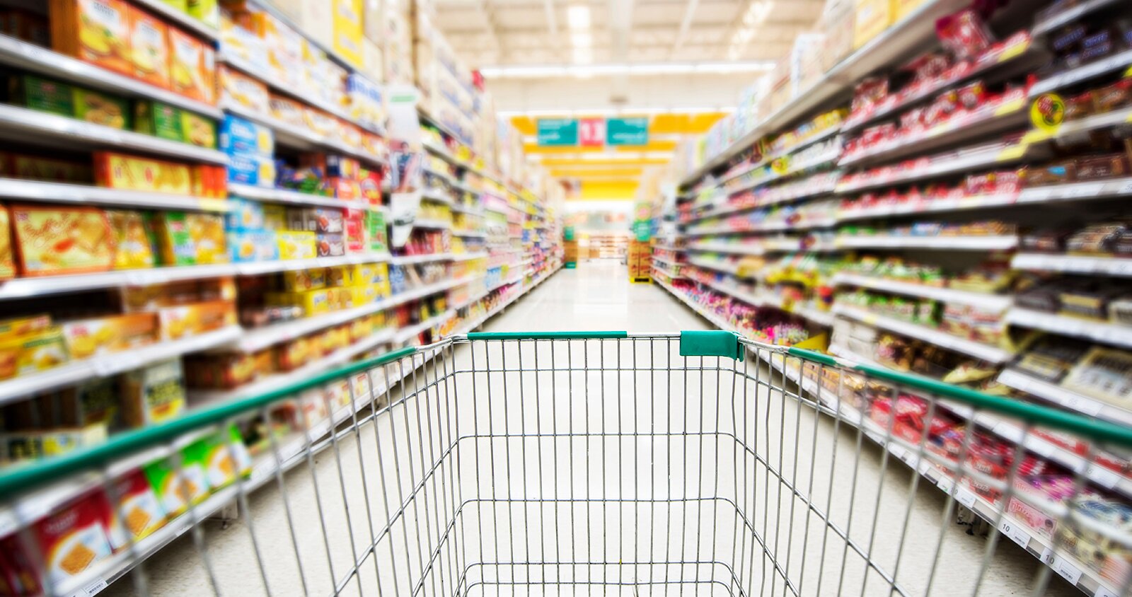 Zdjęcie przedstawia alejkę w supermarkecie. Po prawej i lewej stronie widoczne są rzędy półek wypełnionych różnokolorowymi produktami. Na pierwszym planie pośrodku alejki widoczny jest fragment wózka sklepowego.