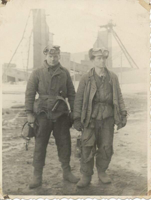 Czarno‑białe zdjęcie przedstawia dwóch mężczyzn z czapkami, na których mają przymocowane latarki. Ubrani są w kurtki, rękawice i grube spodnie. Mężczyzna po prawej trzyma latarenkę. Za mężczyznami znajdują się szyby kopalni.