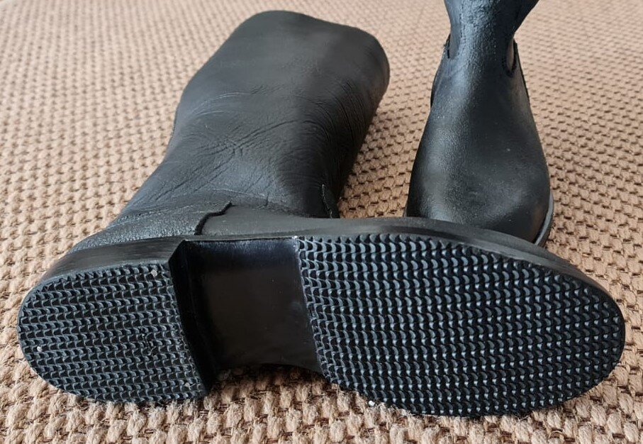 Zdjęcie przedstawia dwa czarne buty z wysoką cholewką i na niskim obcasie. Lewy z nich leży zwrócony podeszwą w stronę obiektywu. Podeszwa jest czarna, ma chropowatą strukturę. Nagranie tożsame z treścią.