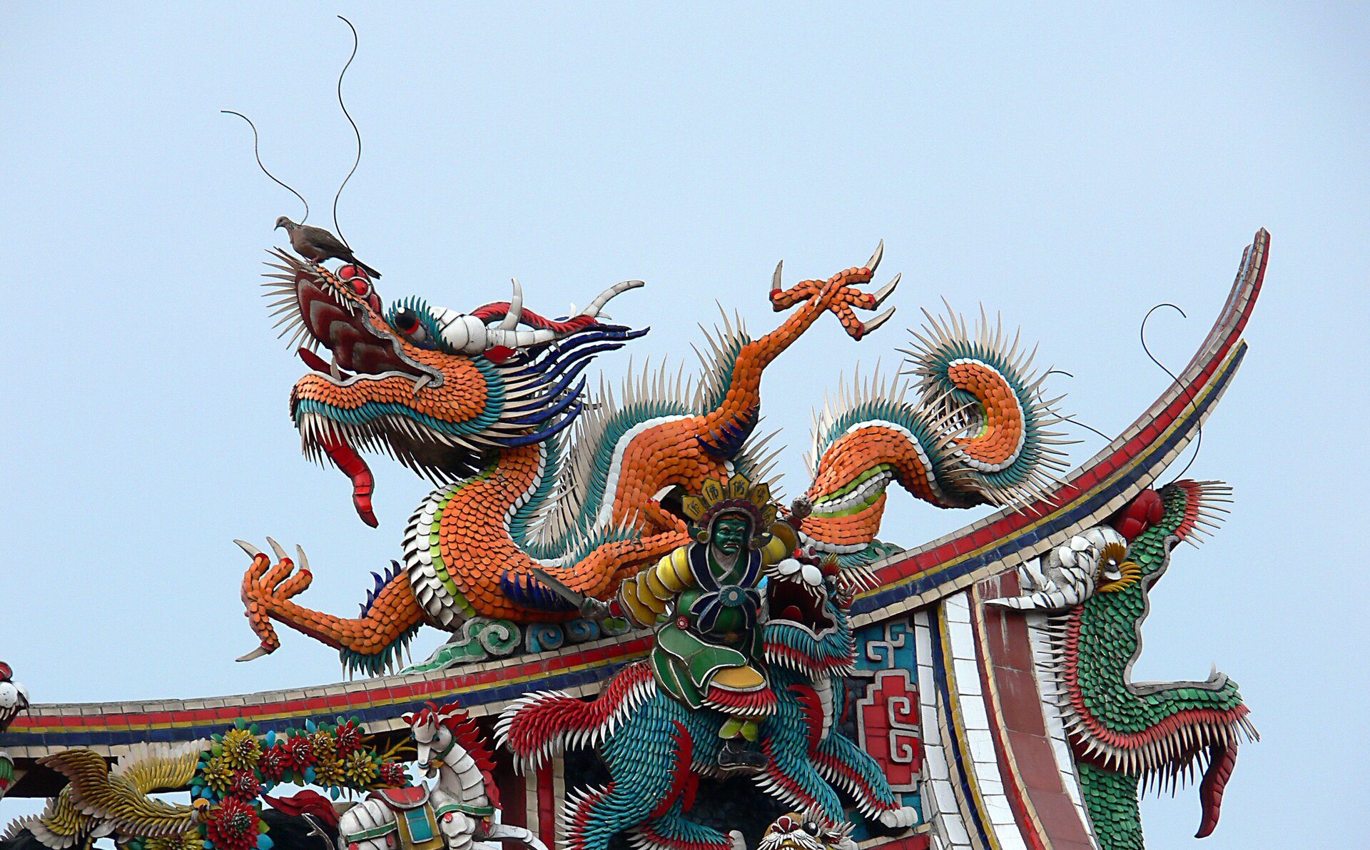 Kliknij, aby powiększyć. Zdjęcie przedstawia smoka na dachu świątyni Longshan w Tajpej. Smok jest niezwykle kolorowy i radosny. Zbudowany jest z mnóstwa kolorowych kamyczków. Ma uniesione łapy i pysk z długimi wąsami. Smok ma otwarty pysk, na którym siedzi żywy ptak. Smok siedzi na budynku, którego fragment widać na zdjęciu. Budynek jest równie kolorowy co smok.