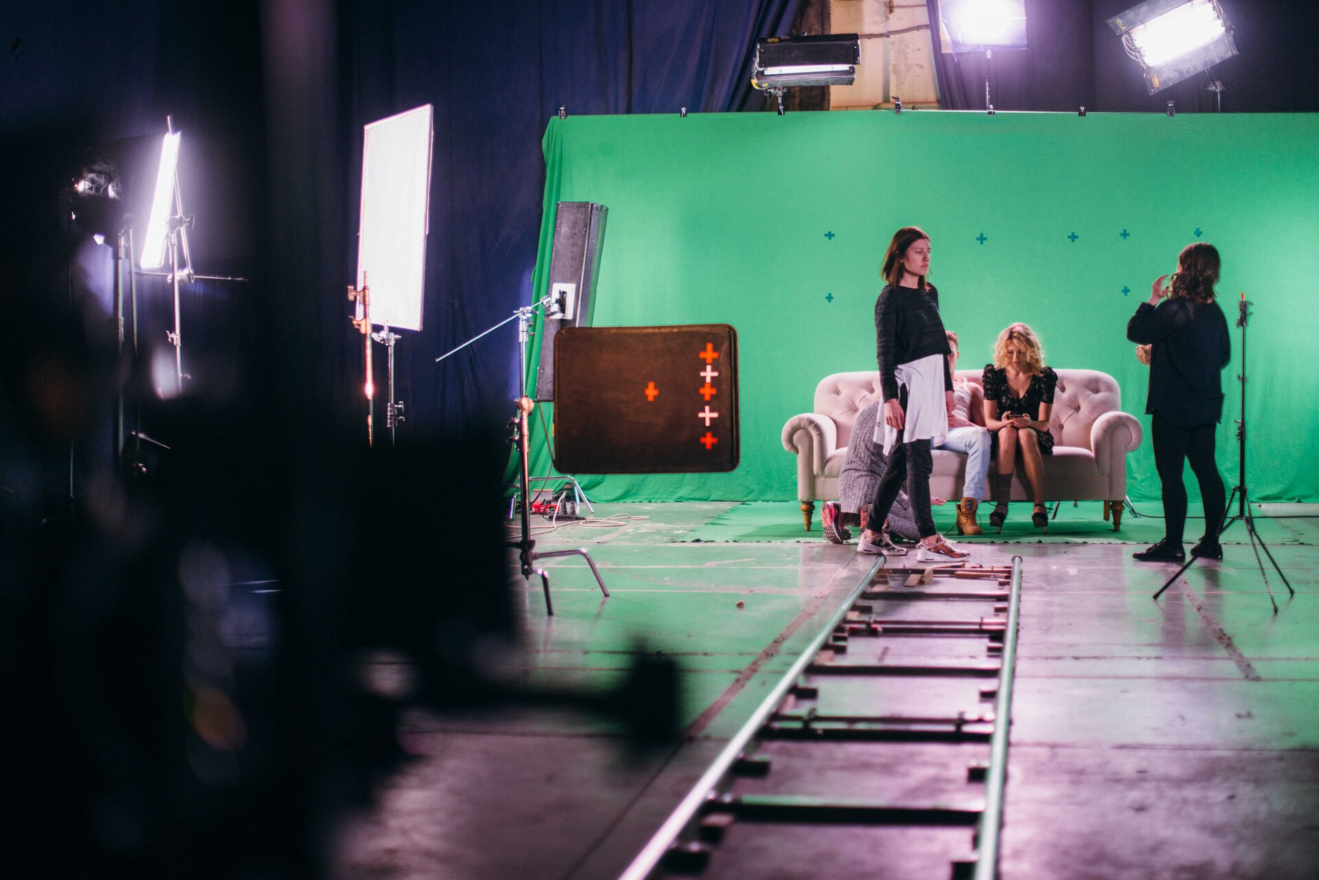 Zdjęcie przedstawia ekipę filmową, sprzęt filmujący i scenografię. W centrum dwoje aktorów siedzących na kanapie na zielonym tle, przy nich trzy osoby z obsługi, przed nimi tory do wózka filmującego, po lewej i nad aktorami reflektory.