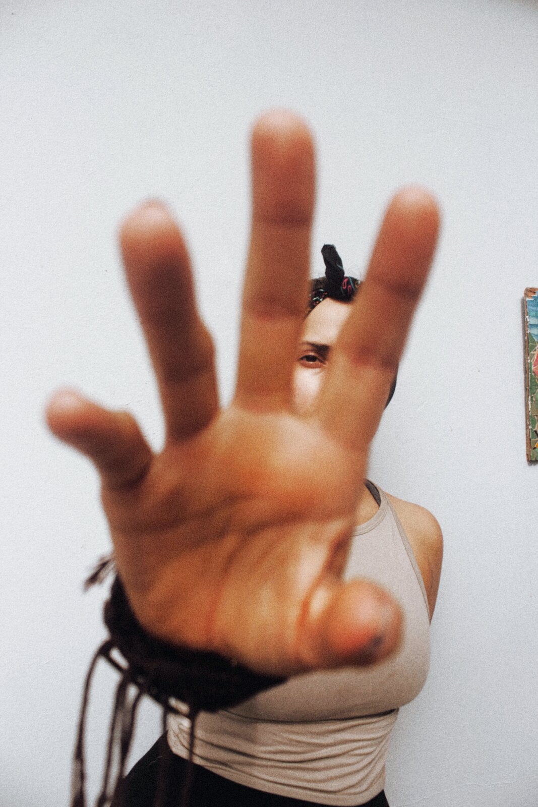 Zdjęcie przedstawia młodą kobietę, w ujęciu od pasa w górę, stojącą na tle białej ściany. Kobieta jest ubrana w beżową koszulkę na ramiączkach, a na głowie ma przewiązaną chustkę. Jej prawa ręka jest wyciągniętą do przodu, a palce dłoni rozczapierzone. Dłoń jest na pierwszym planie i przesłania prawie całą sylwetkę kobiety.