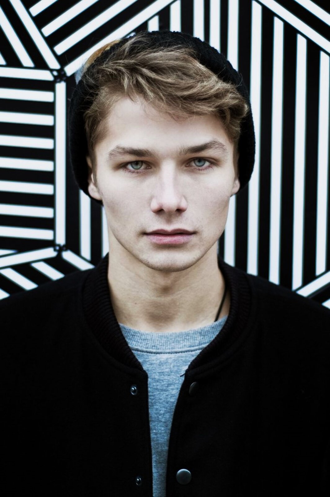 Zdjęcie przedstawia portret chłopca. Chłopak stoi na wprost, patrzy w obiektyw, ma na sobie bluzę,  na głowie czapkę z dzianiny. Za nim ściana w paski, które układają się w różnych kierunkach. 