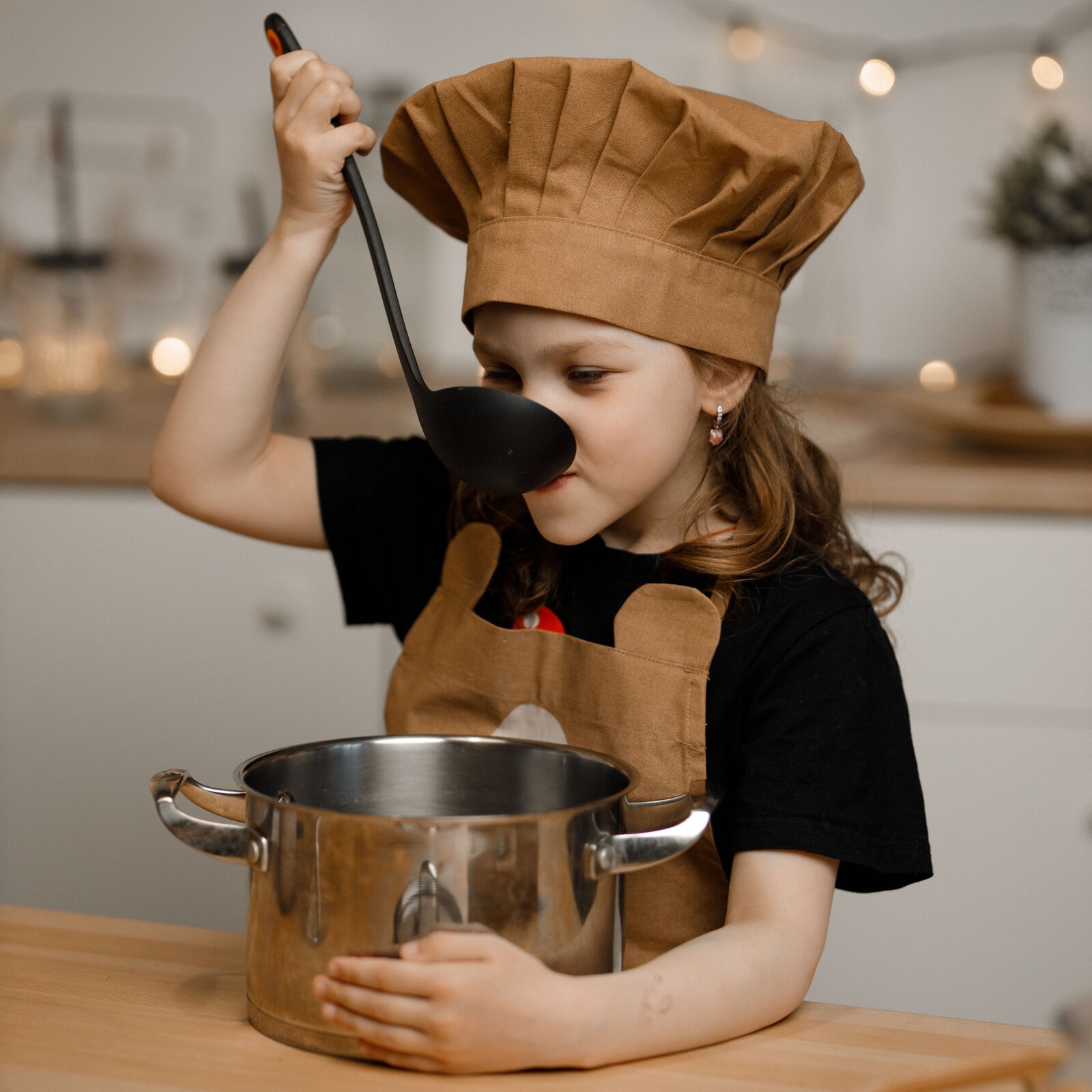 Ilustracja przedstawia dziewczynkę w kucharskiej czapce próbująca zupę.