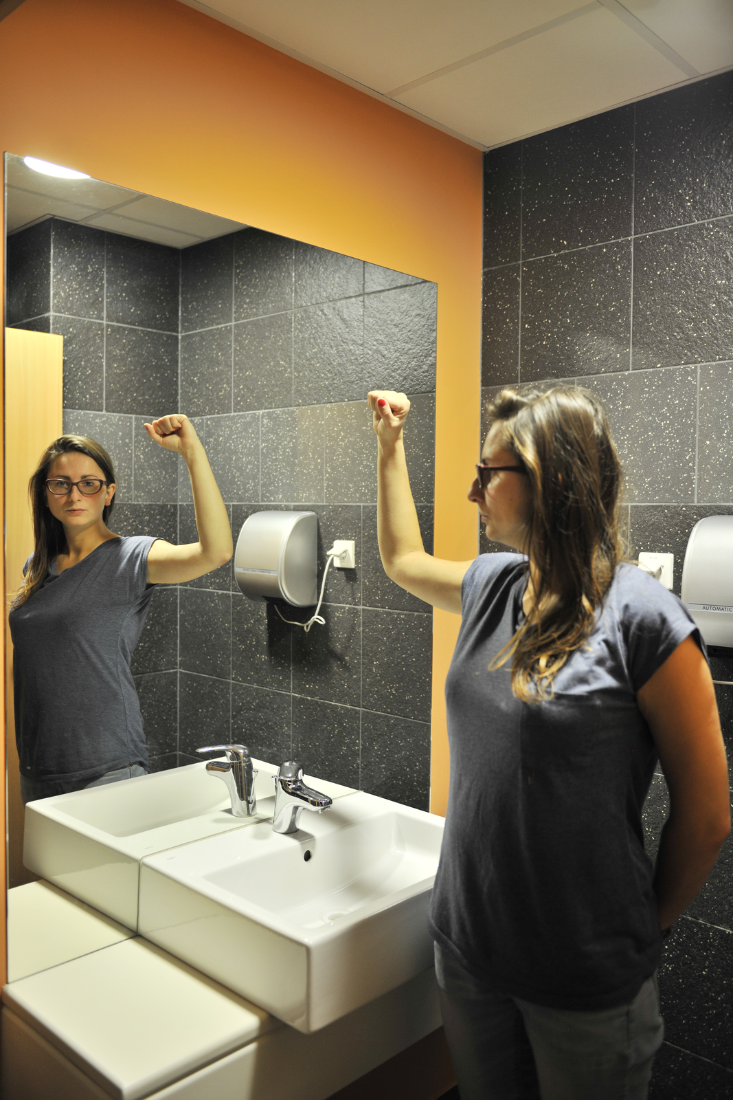 Zdjęcie przedstawia wnętrze łazienki z dużym płaskim lustrem nad białym zlewem. Przed lustrem stoi kobieta  ustawiona bokiem do lustra z uniesioną zgiętą w łokciu ręką. Twarz ma odwróconą od obiektywu aparatu, zwróconą w stronę lustra, ale widać ją dokładnie w odbiciu. Postać kobiety przed lustrem oraz na lustrze jest symetrycznym odbiciem. Tło ciemne.