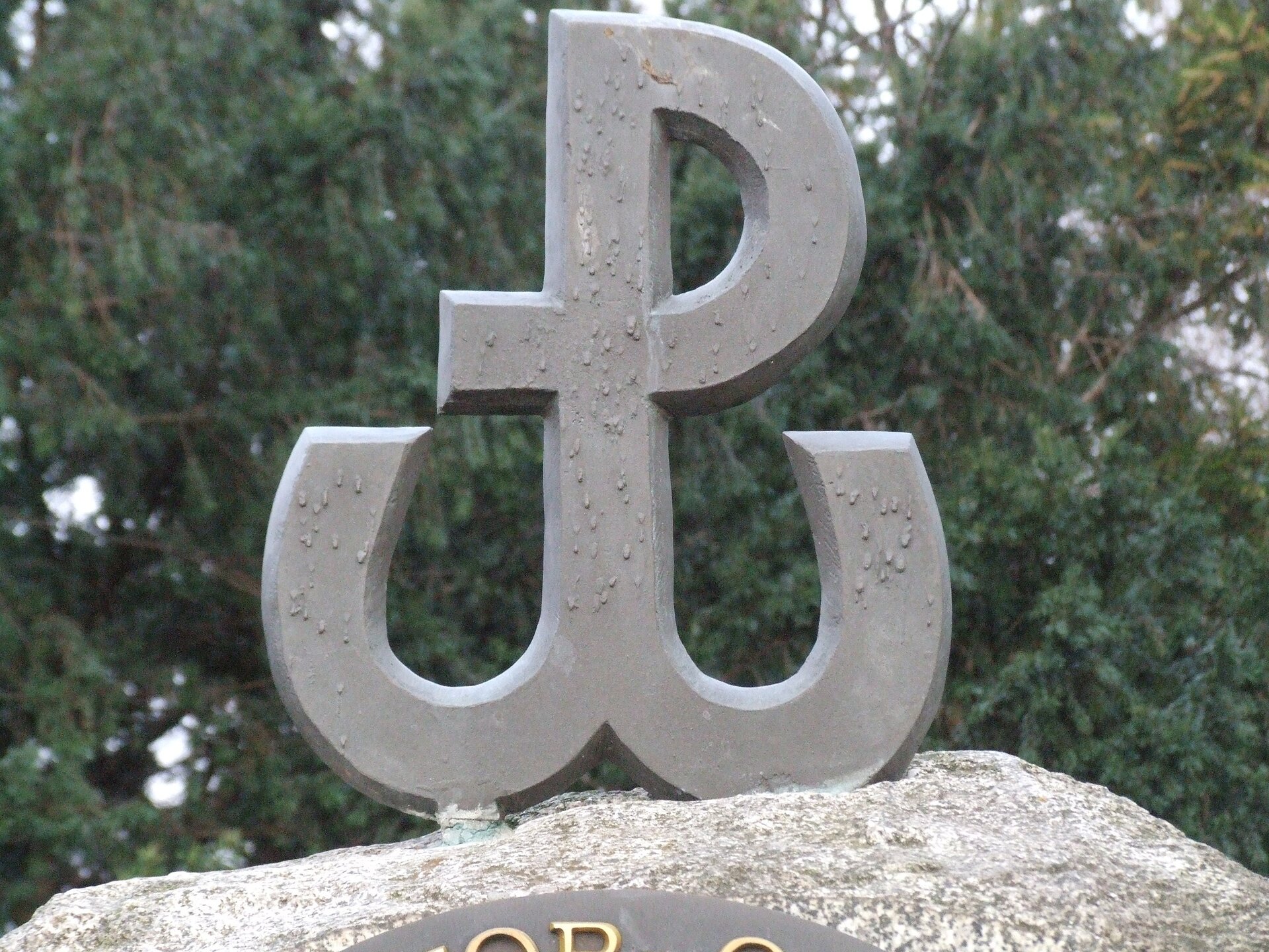 Kolorowa fotografia przedstawia fragment pomnika, którego zwieńczeniem jest litera kształtem przypominająca „P”, u dołu zakończona kotwicą. Na literze widać krople.