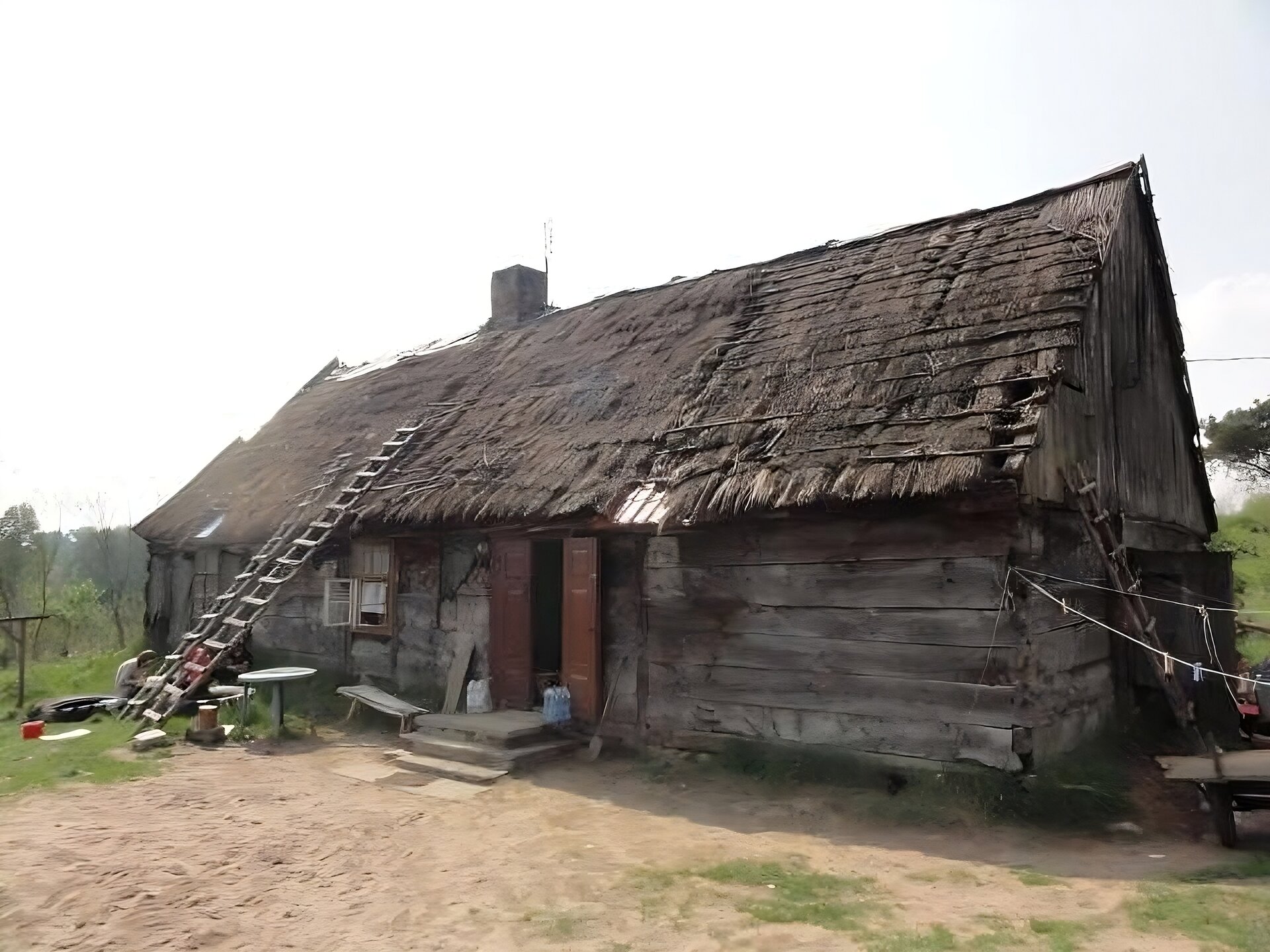 Zdjęcie przedstawia drewnianą, piętrową chatę ze skośnym dachem wykonanym ze słomy. W centralnej części chaty znajdują się podwójne, drewniane drzwi, do których prowadzą dwa ceglane schodki. Z lewej strony budynku stoją dwie drewniane drabiny oparte o dach. Z lewej strony budynku widoczna jest trzecia drabina oparta o bok domu. Przed domem rośnie zielona trawa, a teren najbardziej uczęszczany jest piaskowy. Niebo jest jasne, bezchmurne. Zdjęcie zostało wykonane w świetle dziennym.