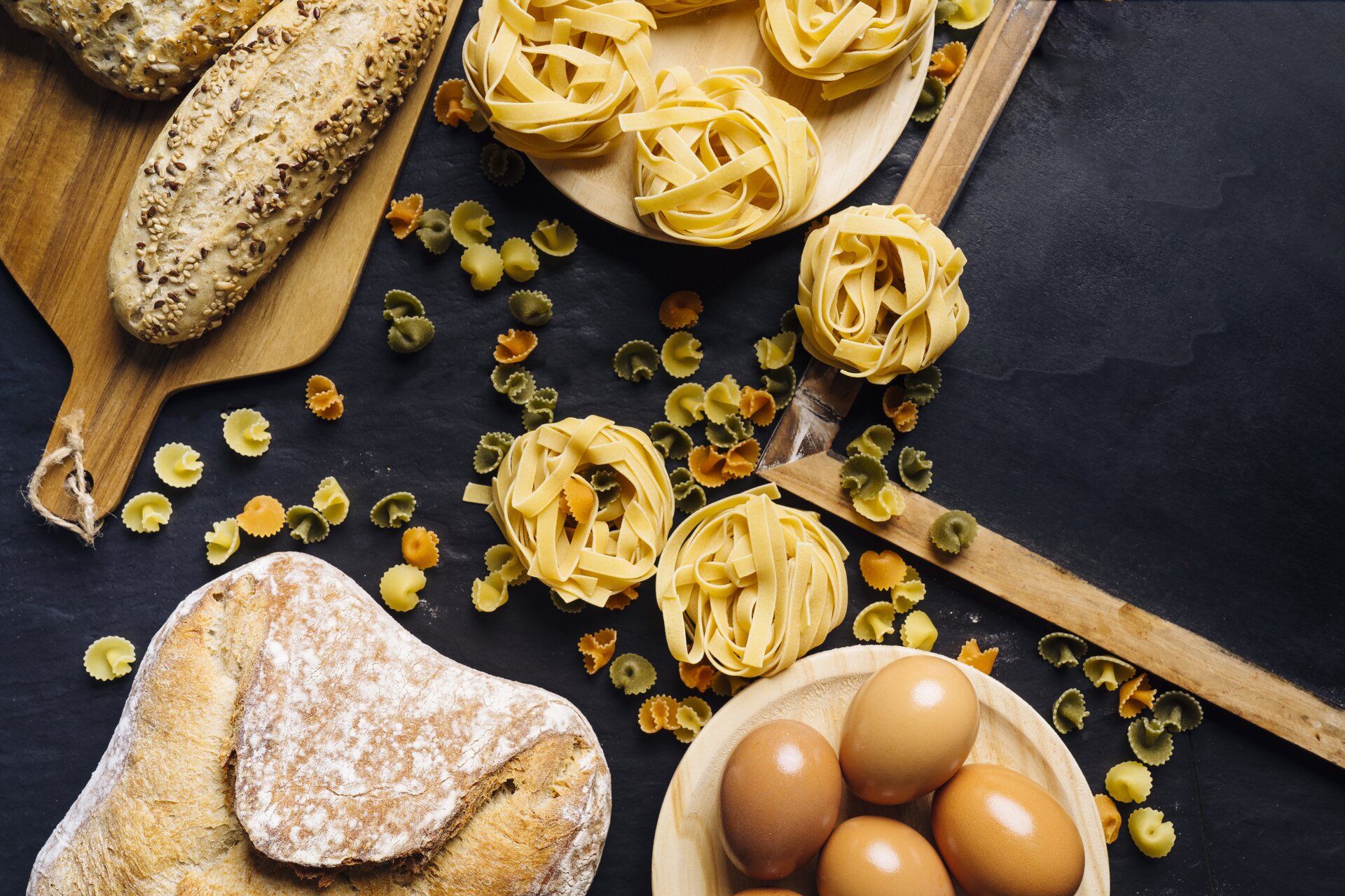 Zdjęcie przedstawia czarny blat, na którym widoczne są: drewniana deska z długą bułką posypaną ziarnami, bochenek chleba, talerzyk z gniazdkami makaronu, trzy gniazdka makaronu, kolorowy makaron muszelki i drewniany talerzyk z czterema jajkami.