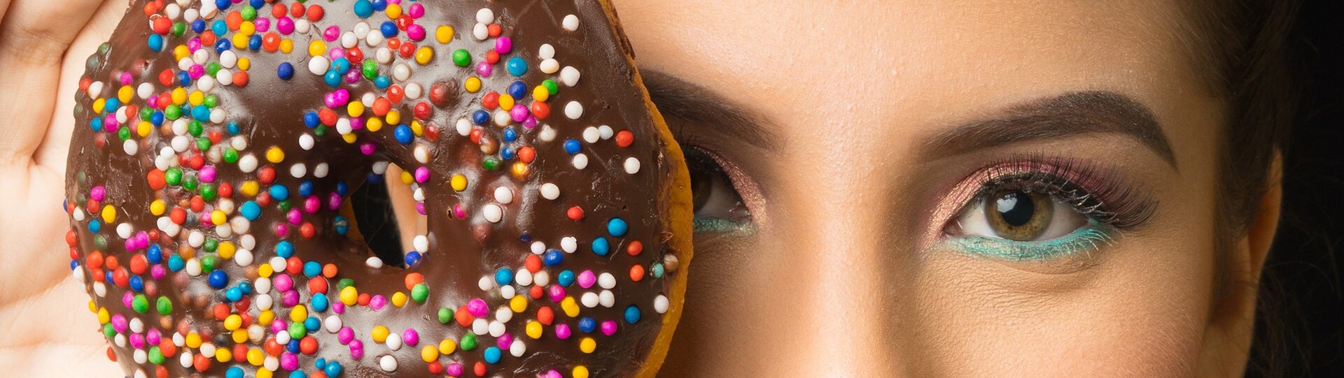 Zdjęcie przedstawia oczy kobiety. Ma nałożony kolorowy makijaż. Jedno z oczu zasłania donatem z czekoladową polewą i kolorową posypką. 
