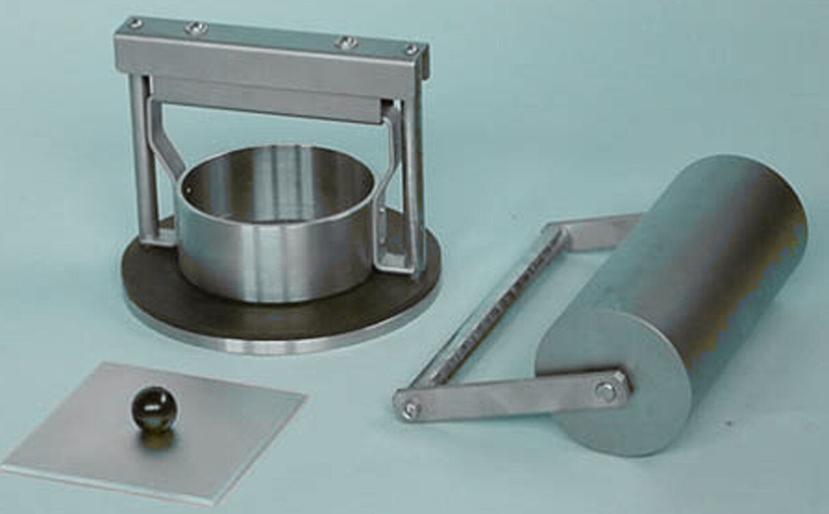 Zdjęcie przedstawia aparat Cobba. Widoczne są trzy elementy. Pierwszy to kwadratowa pokrywka w kolorze srebrnym z czarną okrągłą gałką pośrodku. Drugi to cylindryczny element w kolorze srebrnym, zamocowany na okrągłej podstawie. Na podstawie zamocowany został stelaż z dwóch pionowych rurek z taśmowatymi, łukowato wygiętymi zaczepami, które zostały połączone poziomą poprzeczką. Trzecie element to walec, do którego podstawy i góry przymocowana została rączka.