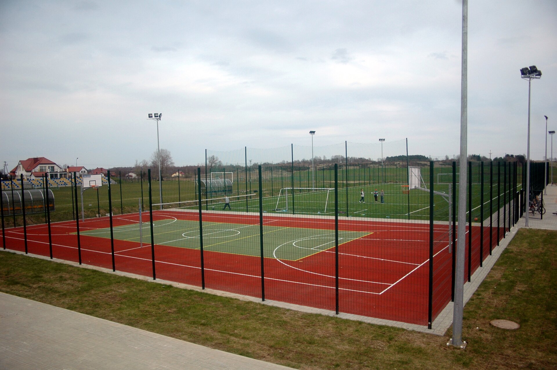 Fotografia przedstawia widok na kompleks sportowy „Orlik” w miejscowości Dygowo. Po lewej stronie znajduje się boisko piłkarskie z trybunami. Na fotografii znajduje się kilka ogrodzonych boisk do gry w piłkę nożną, siatkówkę i koszykówkę ze sztuczną nawierzchnią. Na pierwszym planie widoczne jest wielofunkcyjne boisko do gry między innymi w koszykówkę oraz siatkówkę. Nawierzchnia jest pomarańczowa, a prostokąt wewnątrz – zielony. Widoczne są białe linie dzielące. Za boiskiem do koszykówki, na boisku do gry w piłkę nożną, przy jednej z bramek widać grupę grających. Nad boiskami górują lampy. Fotografia została wykonana w zachmurzony dzień, w tle widać drzewa oraz budynki.