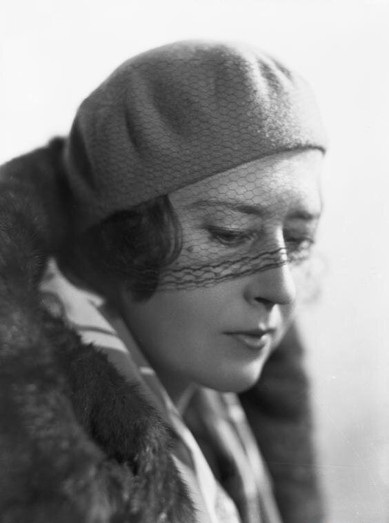 Czarno‑biała fotografia portretowa przedstawia kobietę około 30 roku życia, ubraną w futro. Na głowie ma beret z siatkowaną woalką nasuniętą do połowy twarzy. Pasma włosów wystają spod nakrycia głowy. Kobieta kieruje wzrok w dół, delikatnie się uśmiecha. 
