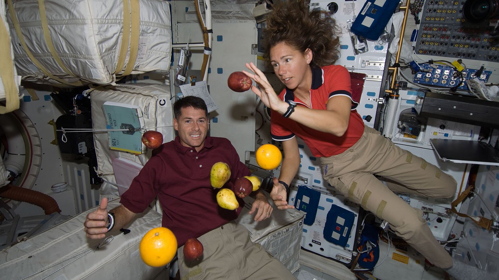 Zdjęcie przedstawia wnętrze statku kosmicznego w trakcie lotu. Widoczne są dwie osoby (kobieta po prawej stronie, a mężczyzna po lewej stronie), które unoszą się w powietrzu. Przed nimi widoczne są również unoszące się owoce: jabłka, gruszki, pomarańcza i grapefruit. W tle widoczna jest aparatura oraz różnego rodzaju pojemniki.