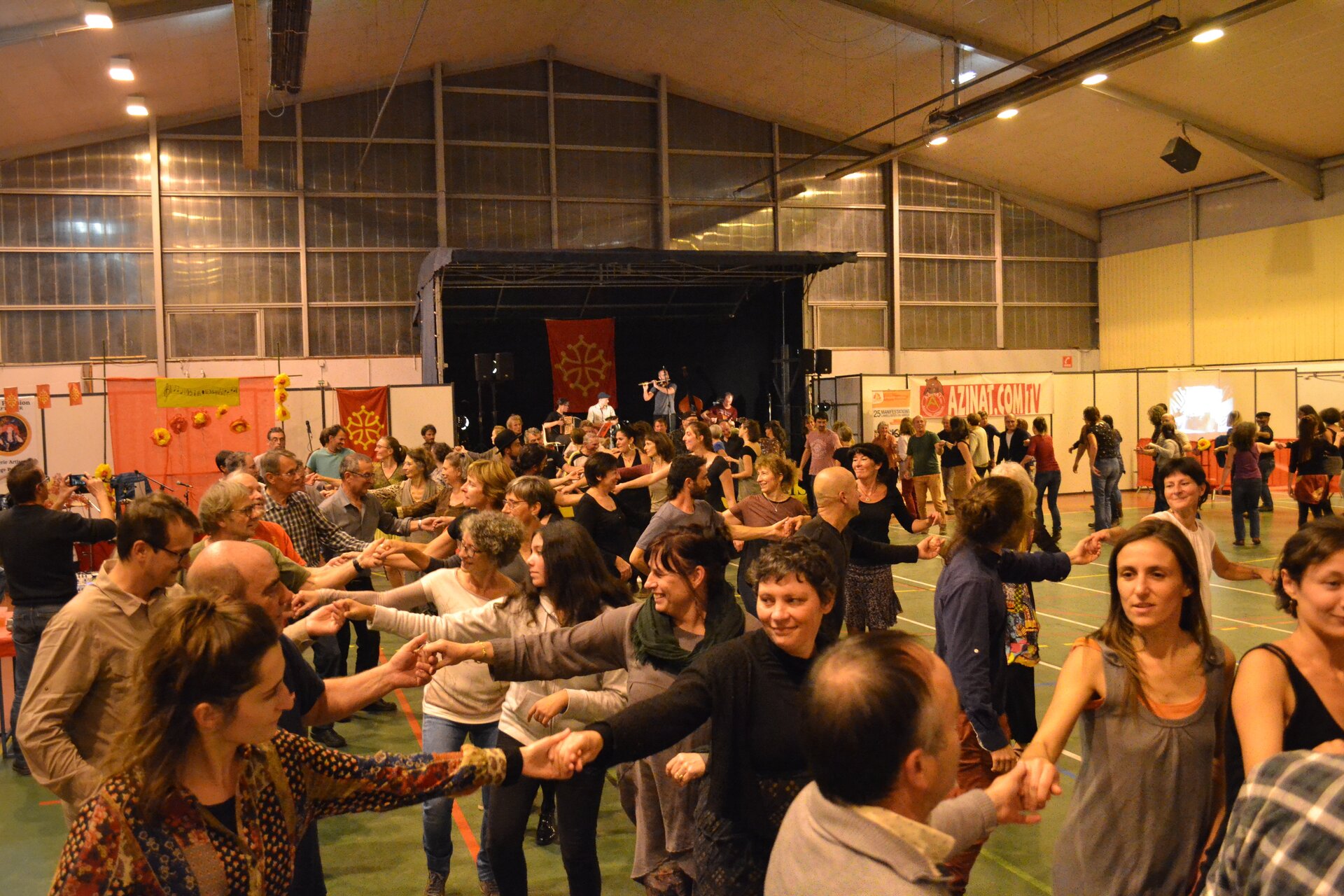 Fotografia prezentuje grupę ludzi tańczących „Belgijkę” w czasie balu, który odbywa się na hali. Pomieszczenie jest obszerne, z belkami przytrzymującymi konstrukcję ściany uchwyconej na wprost. Podłoże jest zielone z obecnymi gdzieniegdzie pomarańczowymi liniami. Obecni w pomieszczeniu ludzie dobrani są w pary, trzymając się za ręce. Obecne są pary męsko‑damskie oraz jednopłciowe. Są to ludzie w różnym wieku. Z tyłu uchwycono scenę oraz ludzi grających na instrumentach. Na scenie przymocowano plakat – czerwony prostokąt ze złotym kształtem przypominającym krzyż. 
