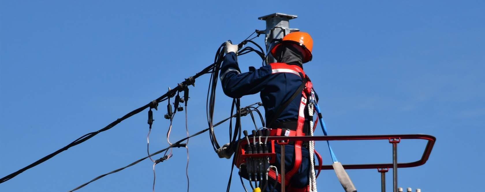 Zdjęcie przedstawia elektryka podczas pracy na wysokości w trakcie naprawy linii wysokiego napięcia. Mężczyzna pracuje z podnośnika, ma na sobie pomarańczowy kask, białe rękawice ochronne, niebieską kurtkę roboczą oraz biało czerwone szelki bezpieczeństwa. Szelki przypięte są do podnośnika dwiema linkami w celu zabezpieczenia elektryka przed upadkiem.