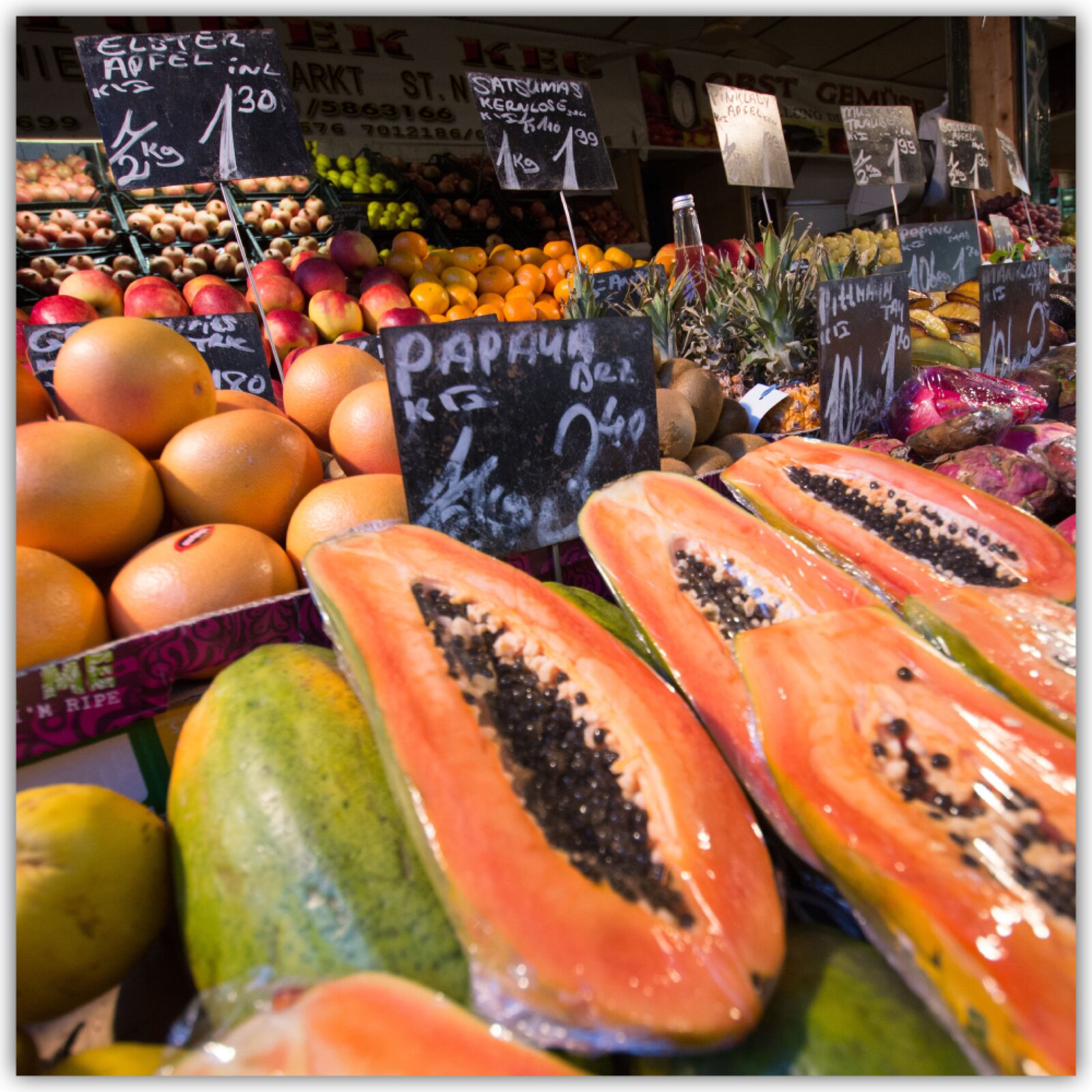 Grafika przedstawia stoisko z owocami. Są na nim między innymi papaje, grejpfruty, jabłka, ananasy.