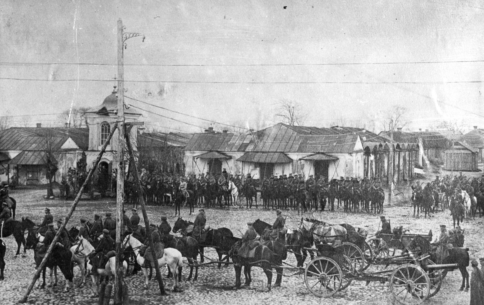 Zdjęcie przedstawia oddziały wojska polskiego. Na pierwszym planie są żołnierze w mundurach i czapkach na koniach wraz z powozami. Za nimi jest pusty plac, na którym na koniu jest dowódca. Za nim jest dalsza część oddziału. W tle są zabudowania rynku.