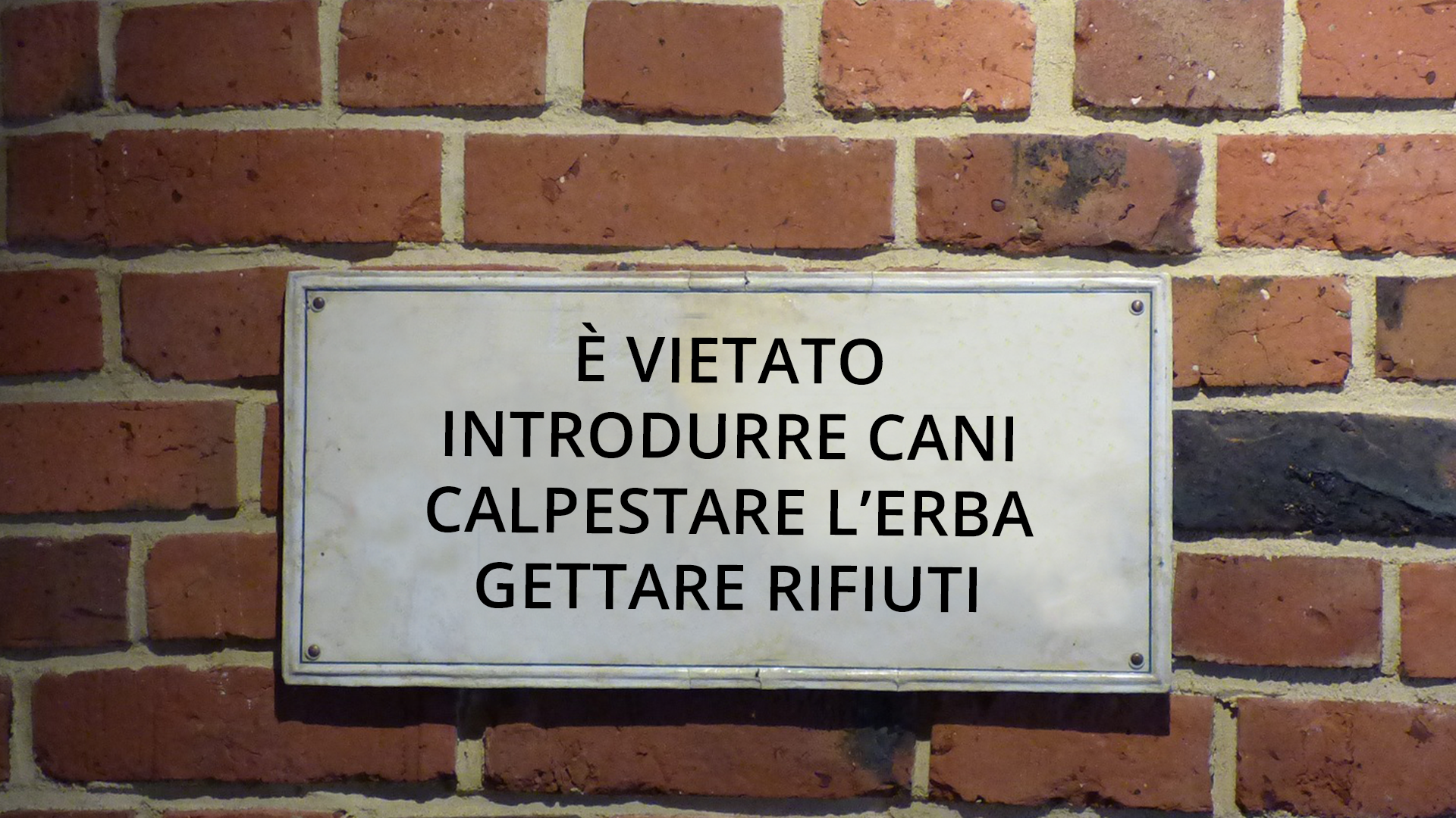 Zdjęcie przedstawia tabliczkę umieszczoną na ścianie z cegły. Na tabliczce znajduje się napis: È vietato: introdurre cani calpestare l’erba gettare rifiuti.