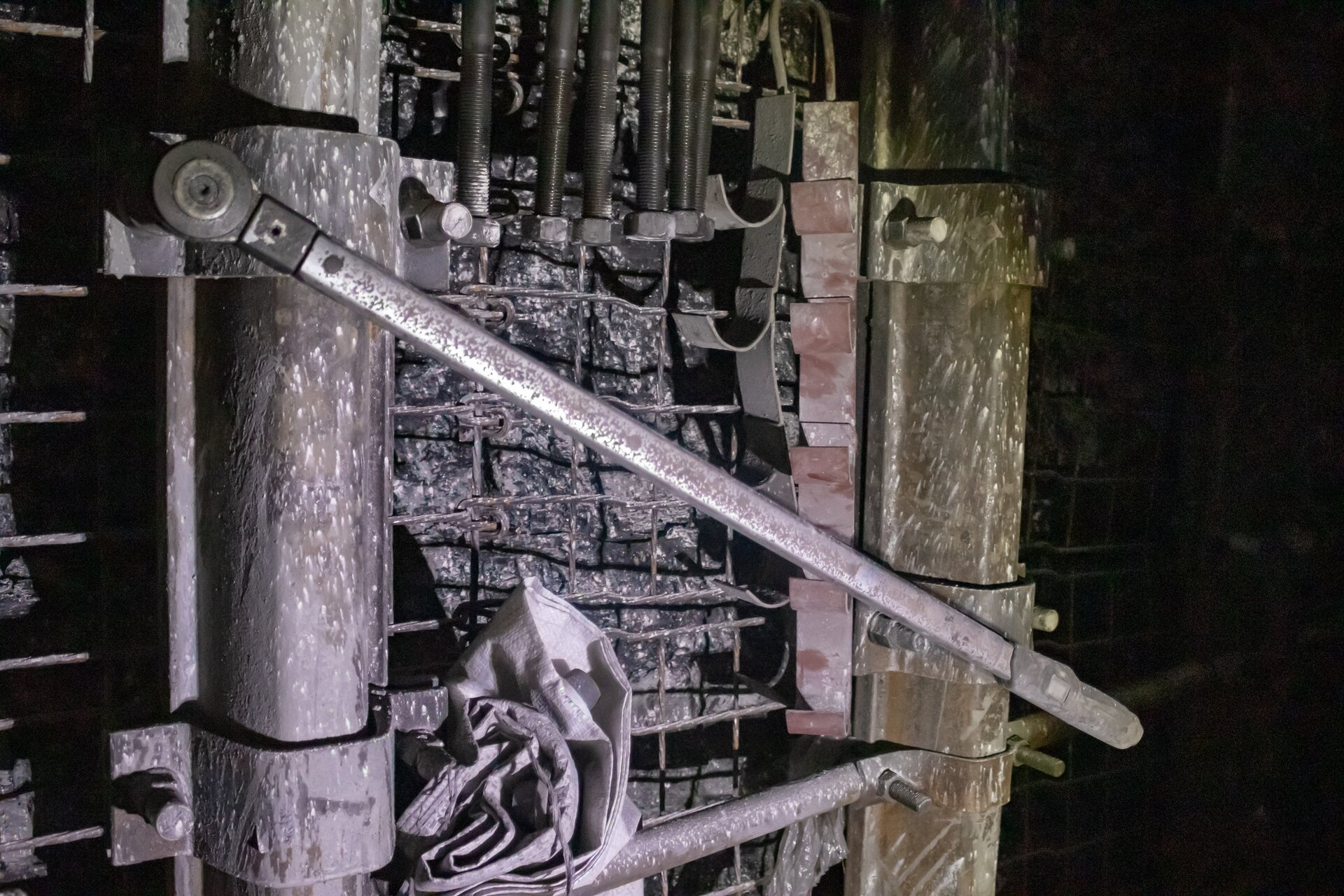 Na zdjęciu widać klucz dynamometryczny do skręcania zamków. Klucz ma kształt podłużnego cienkiego prostokąta o kształcie zbliżonym do dźwigni. W lewej części klucz jest przymocowany do elementu o kształcie niewielkiego cylindra.W tle widać fragment zabezpieczonego ociosu.