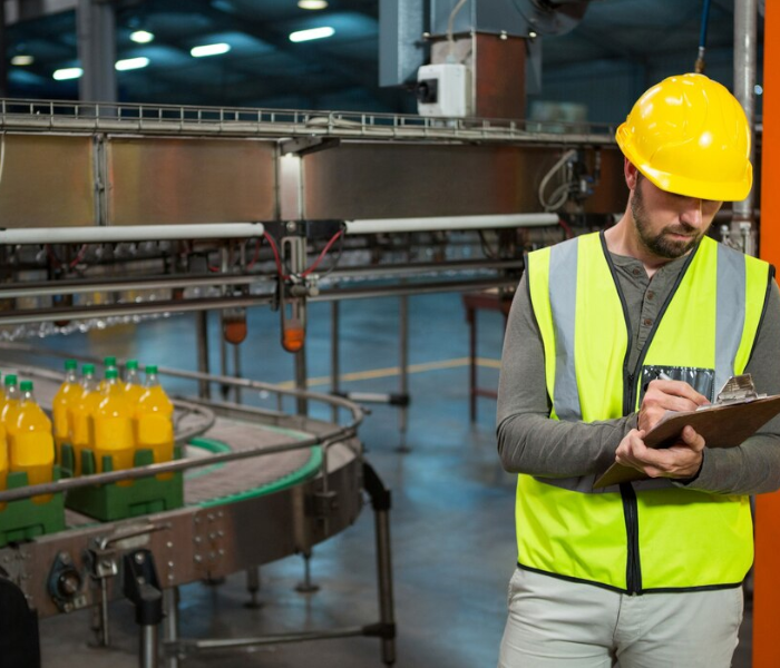 Zdjęcie drugie, podpisane Transportowanie gotowych produktów z linii produkcyjnej do magazynu. Na zdjęciu widać mężczyznę ubranego w żółtą kamizelkę odblaskową i żółty kask. Stoi na hali produkcyjnej przy taśmociągu, na którym widoczne są zgrzewki soku pomarańczowego. Mężczyzna w dłoniach trzyma podkładkę z dokumentacją, w której coś zaznacza.