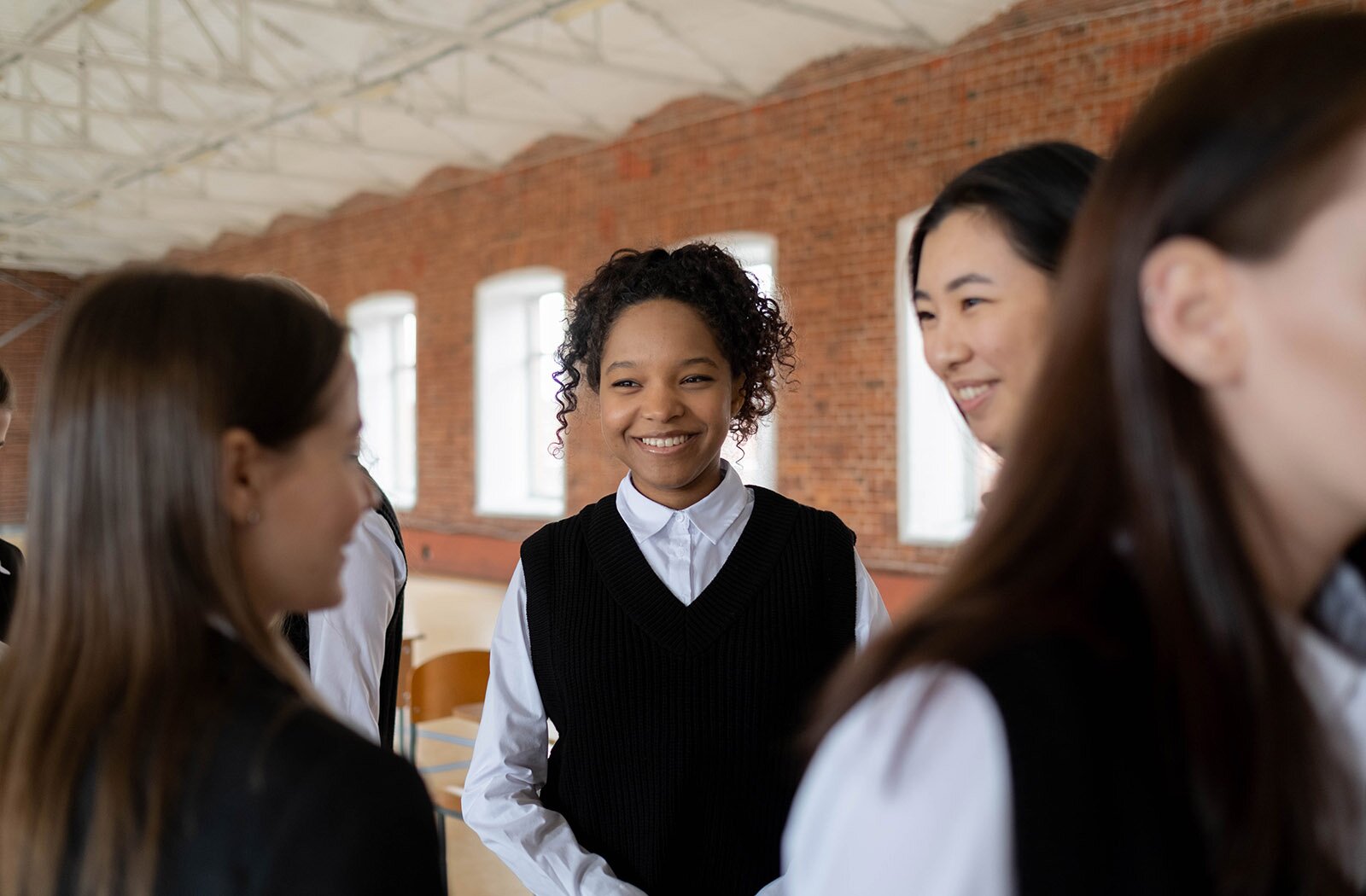 Zdjęcie przedstawia nastolatki w pomieszczeniu. Ubrane są w białe koszule z długim rękawem i czarne kamizelki. Trzy dziewczyny stoją w grupie, rozmawiają i uśmiechają się. Na pierwszym planie widoczny jest fragment nastolatki zwróconej w prawo.