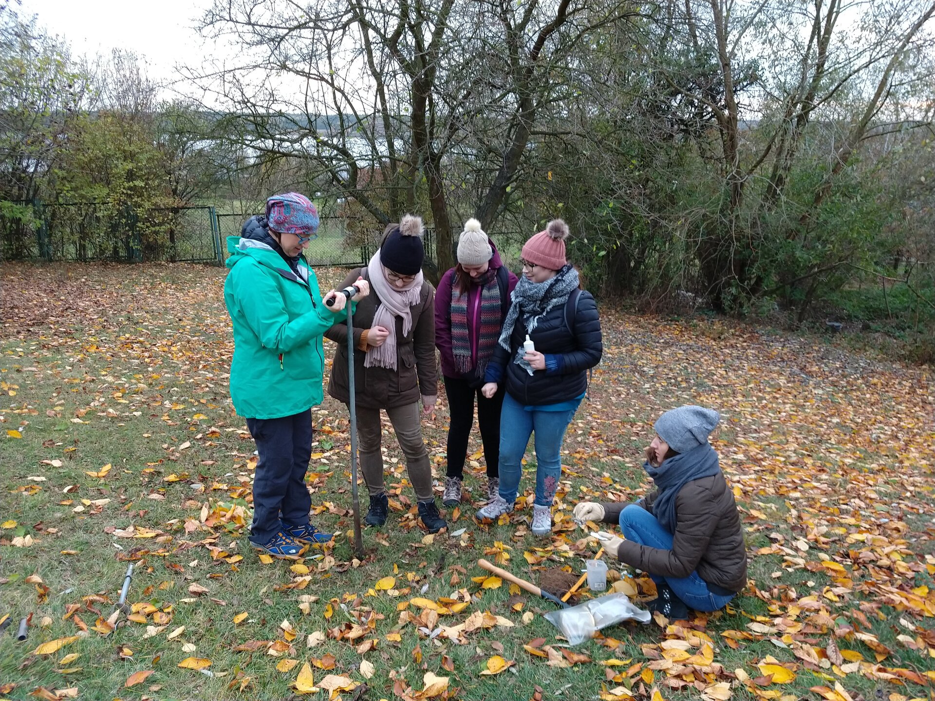 Na zdjęciu grupa studentów gromadzonych jesienią na trawie pokrytej liśćmi. Stoją obok prowadzącej, która pobiera próbkę ziemi za pomocą długiej, pionowej rury z poziomym uchwytem na górze.