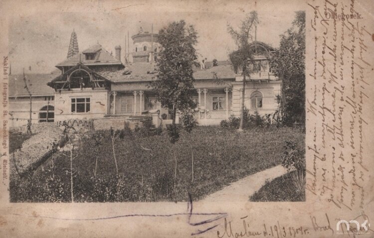 Fotografia przedstawia kartkę pocztową ze zdjęciem prezentującym pałac. Na pierwszym planie znajduje się trawnik, młode drzewa i ścieżka wiodąca w prawą stronę. Pałac jest w trakcie rozbudowy. Ściany są przysłonięte drewnianymi rusztowaniami. W otworach okiennych nie ma szyb. Zadaszenie piętra również nie jest dokończone. Okrągła wieża znajdująca się w głębi sięga drugiego piętra. Wokół niej piętrzy się rusztowanie.