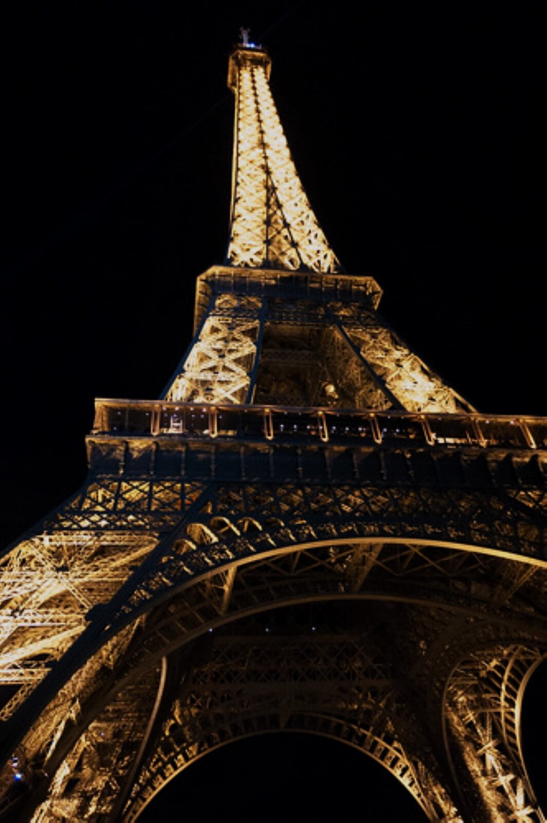 Fotografia przedstawia Wieżę Eiffla w Paryżu. Zdjęcie zostało wykonane w nocy, z perspektywy żabiej, czyli od dołu. Oświetlona konstrukcja wieży połyskuje majestatycznie w mroku; jej dolna część wydaje się ogromna, zwęża się, biegnąc ku górze.  Odnosi się wrażenie jakby sięgała nieba.