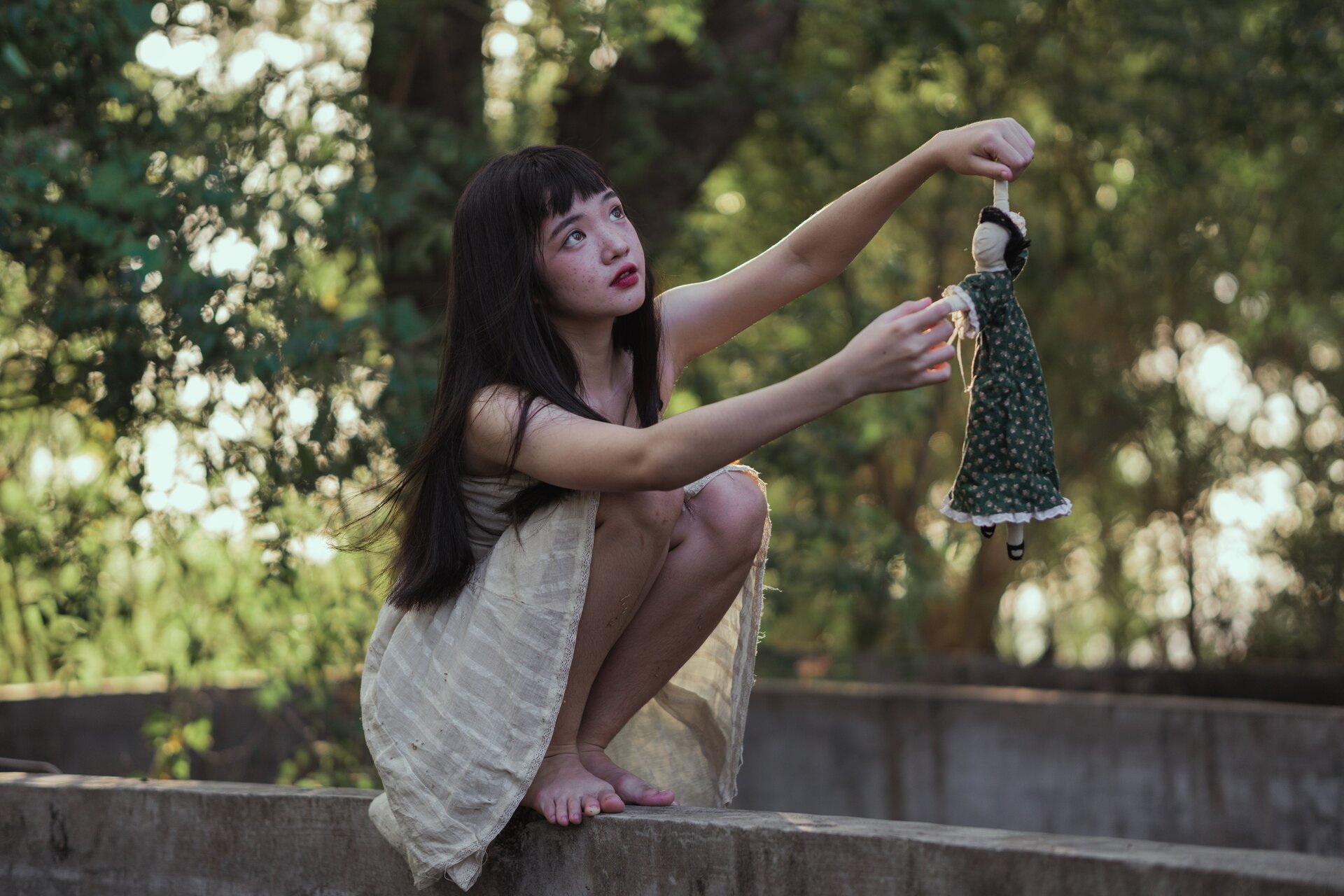 Zdjęcie przedstawia młodą dziewczynę, która siedzi na murku. W ręce trzyma szmacianą lalkę.